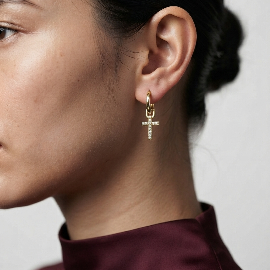 A close-up of a model's ear wearing a small gold hoop earring with a dangling cross charm embellished with clear stones, paired with a dark burgundy top against a light gray background.