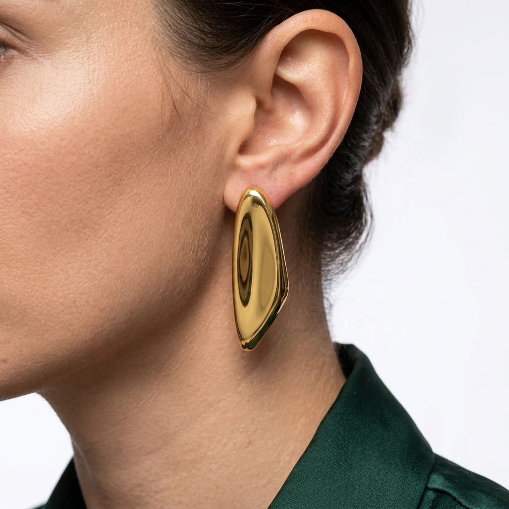 A close-up side profile of a woman wearing a large, polished gold geometric drop earring. The earring has a smooth, reflective metallic surface with a circular cutout detail in the center Contemporary stainless steel statement stud earrings in a high polish gold finish designed with an elegant elongated sculptural shape by Civaro