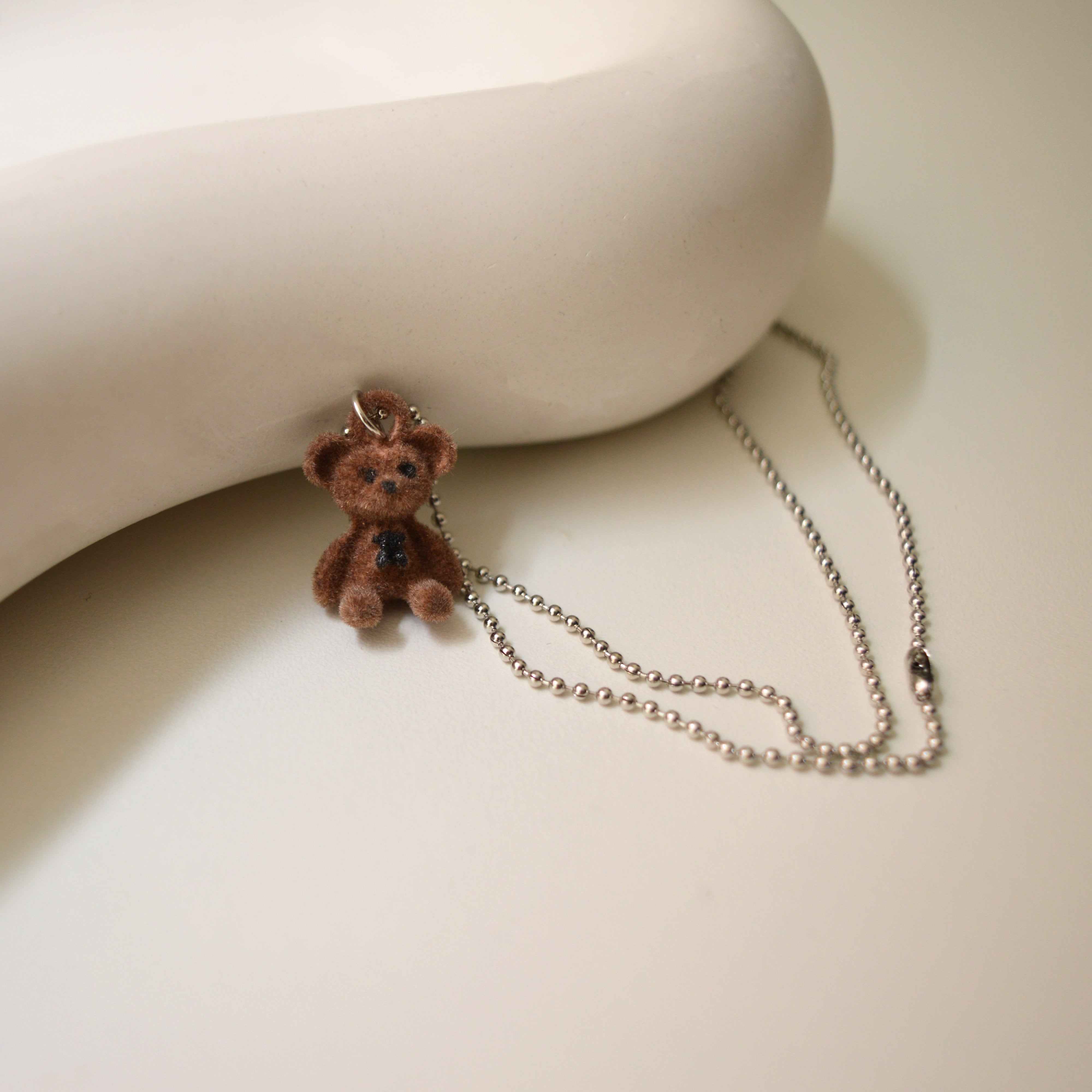 A silver chain necklace featuring a small brown teddy bear pendant with a dark blue bow tie, displayed on a warm off-white surface next to a matching curved display stand by Civaro
