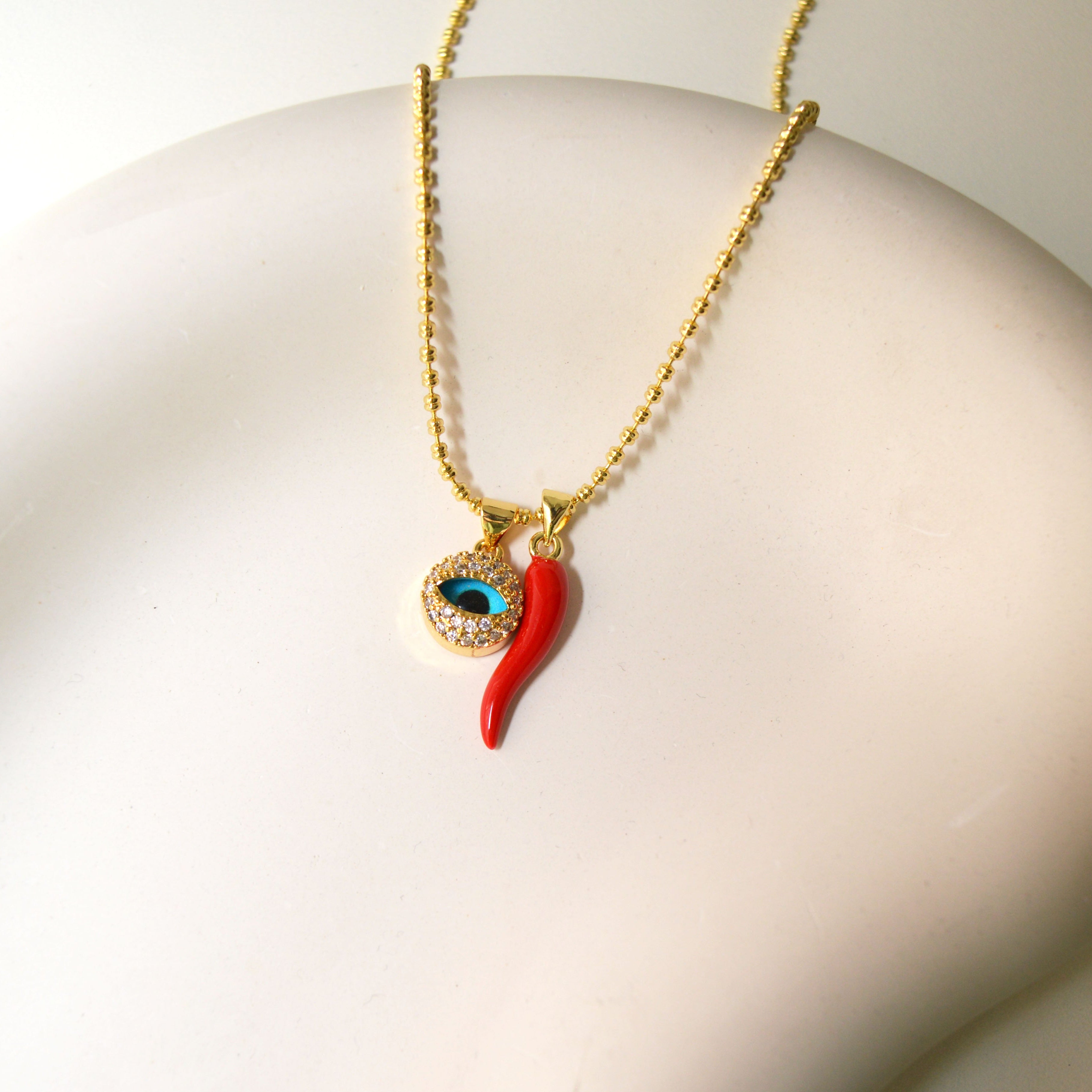 A gold chain necklace with a pendant featuring a red chili pepper and an evil eye design, displayed on a white mannequin bust.