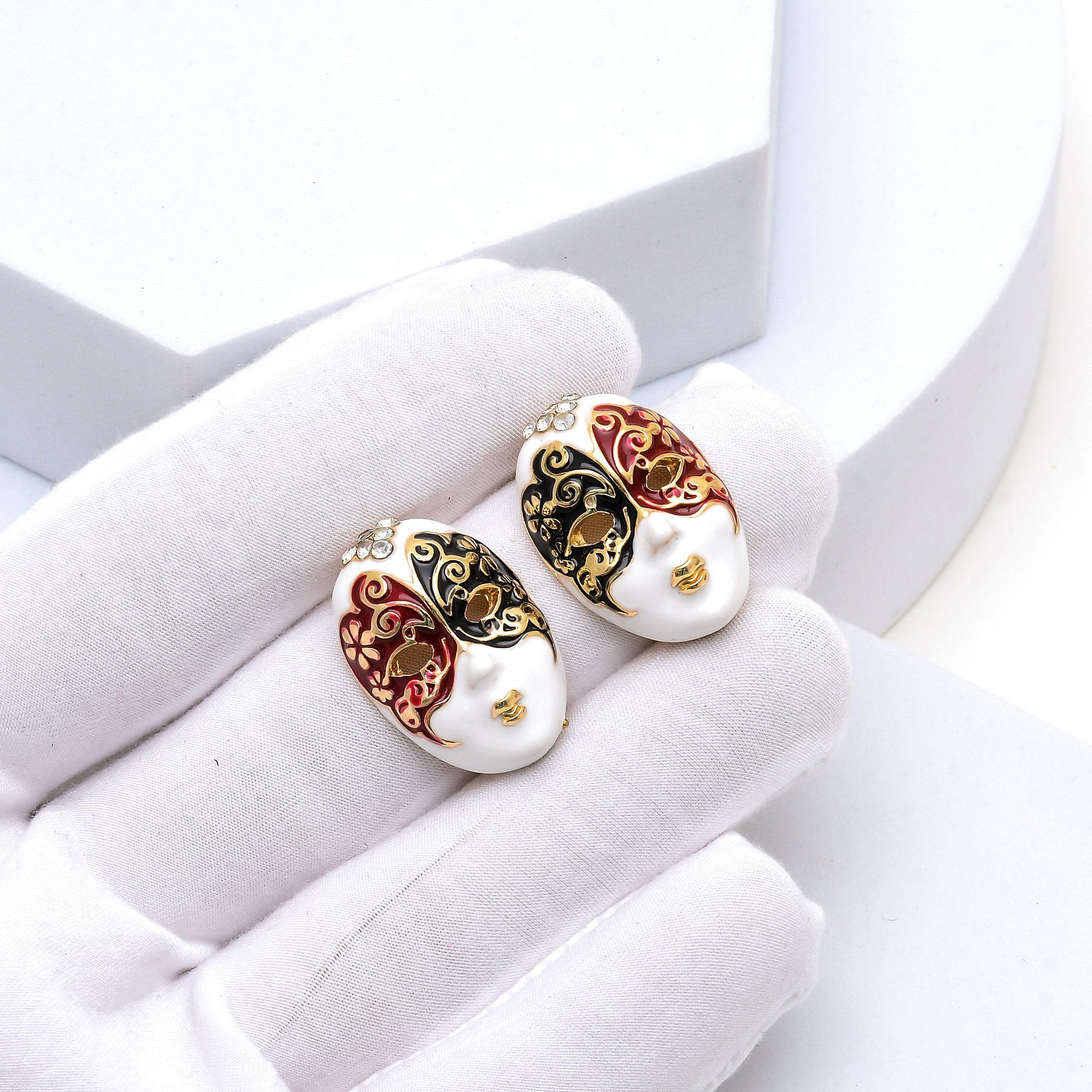 Close up of the shimmering crystal crown and smooth enamel detailing on the Masque Surrealist earrings held in hand