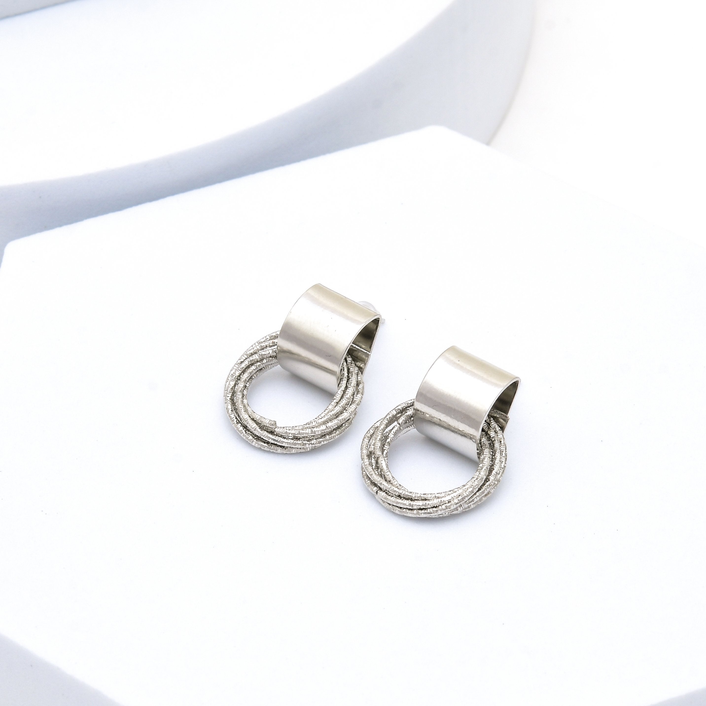 A pair of silver-toned drop earrings with a textured circular wire design and a smooth cylindrical cap, displayed on a clean white surface by civaro
