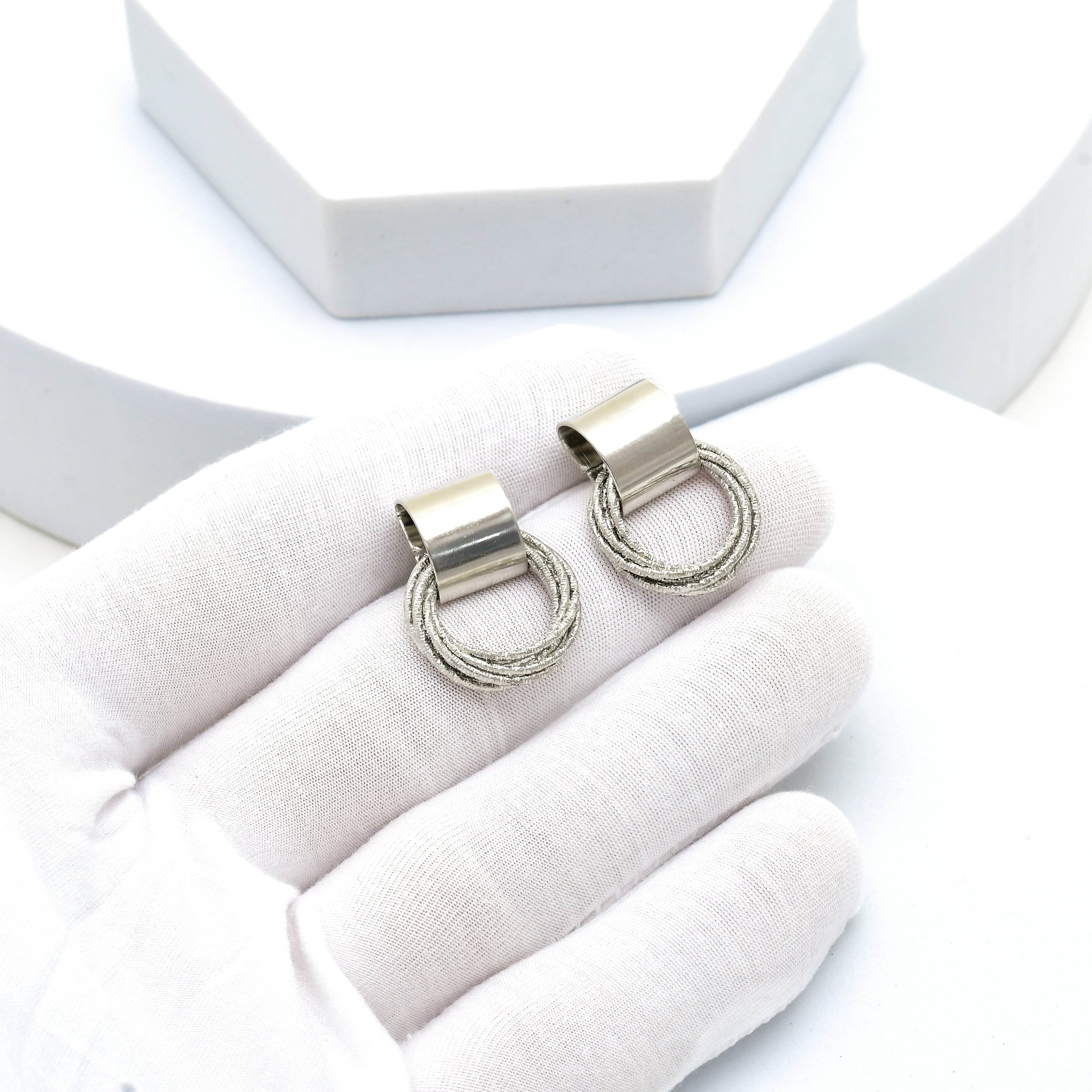 A pair of silver-toned metal ring charms with a textured rope-style lower loop, displayed on a white fabric glove over a white background with a white geometric prop by civaro