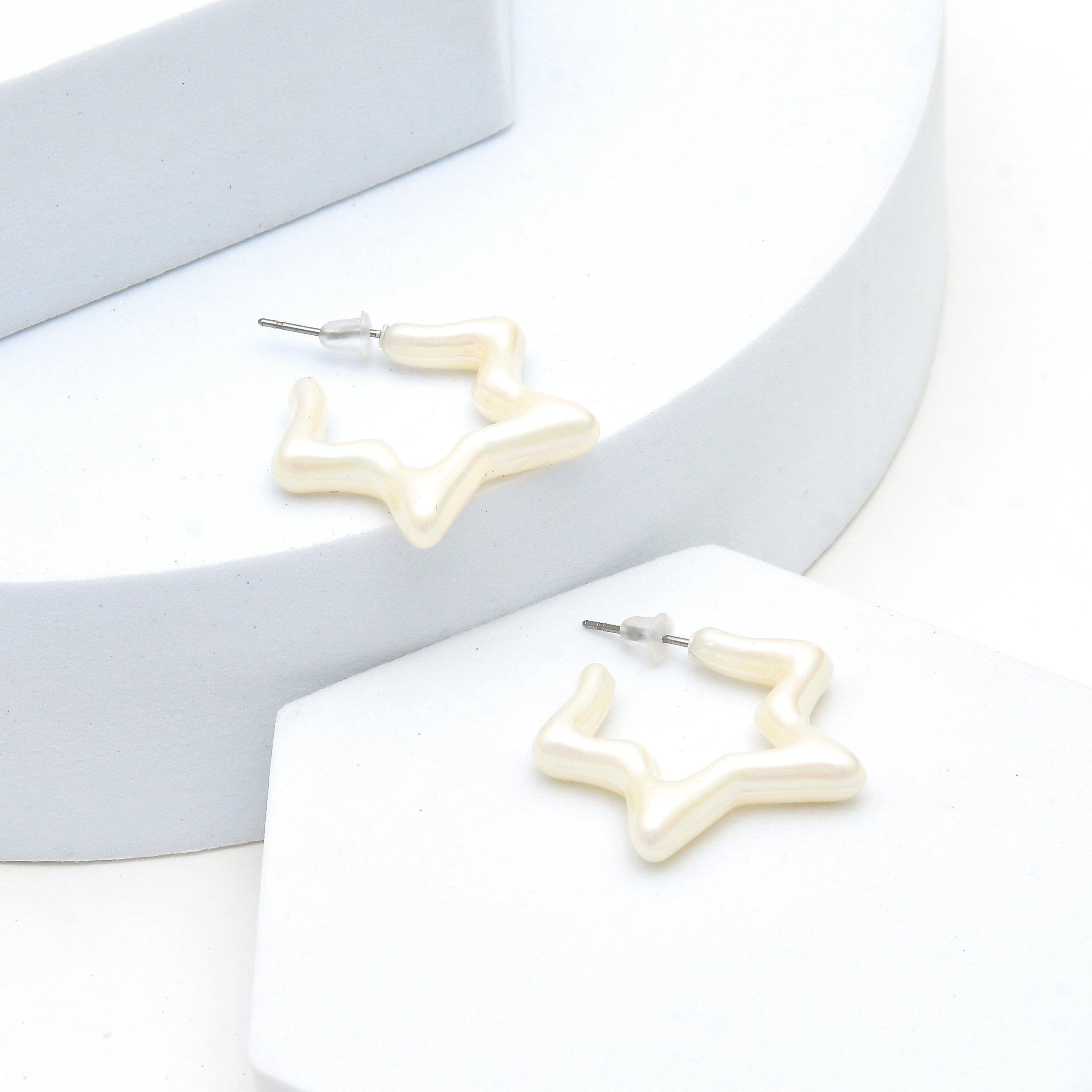 A pair of irregular, organic-shaped hoop earrings with a glossy cream or off-white finish, displayed on white geometric display blocks, Lightweight white statement earrings with pearl inspired finish by Civaro