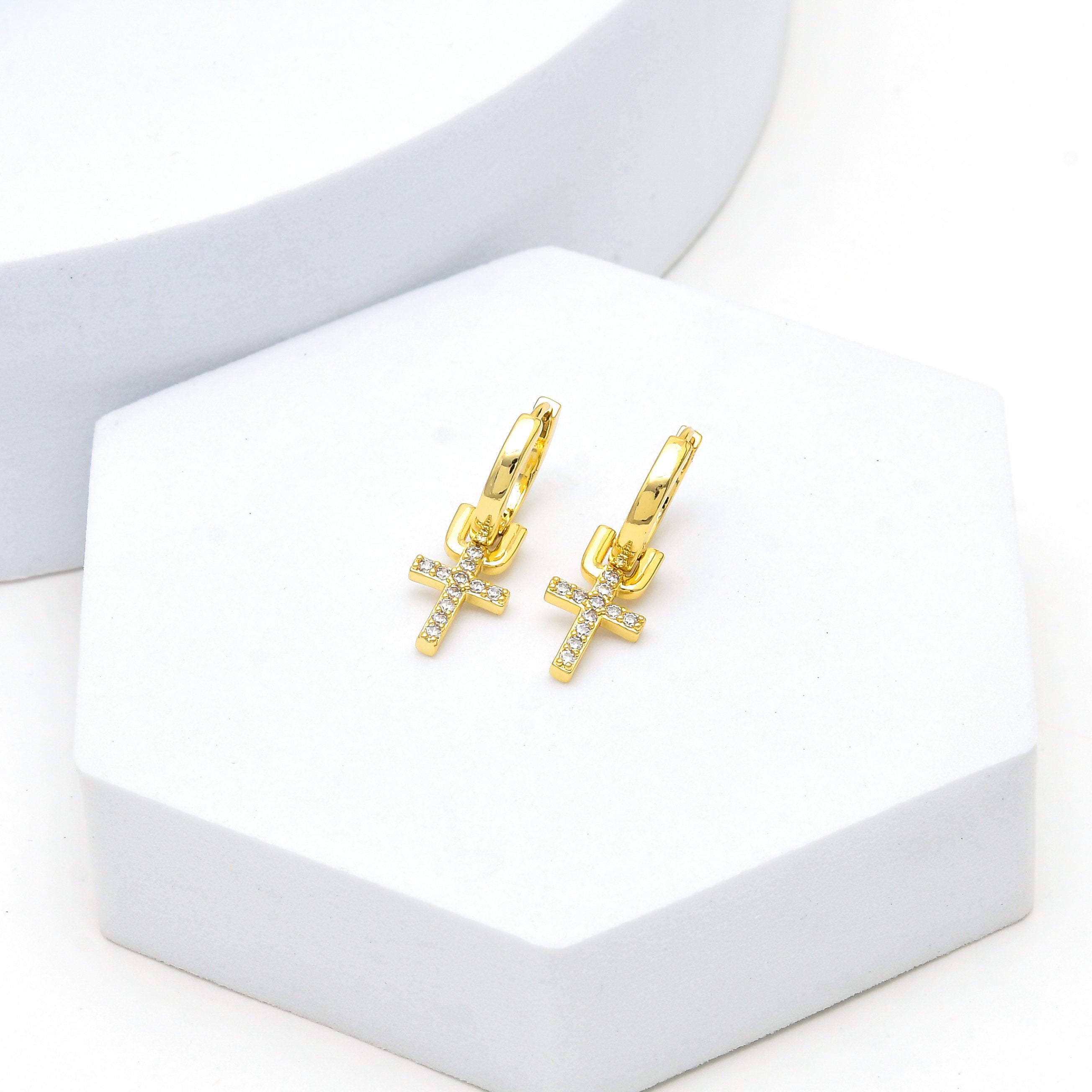 A pair of gold hoop earrings with small cross charms set with clear stones, displayed on a white hexagonal stand by Civaro