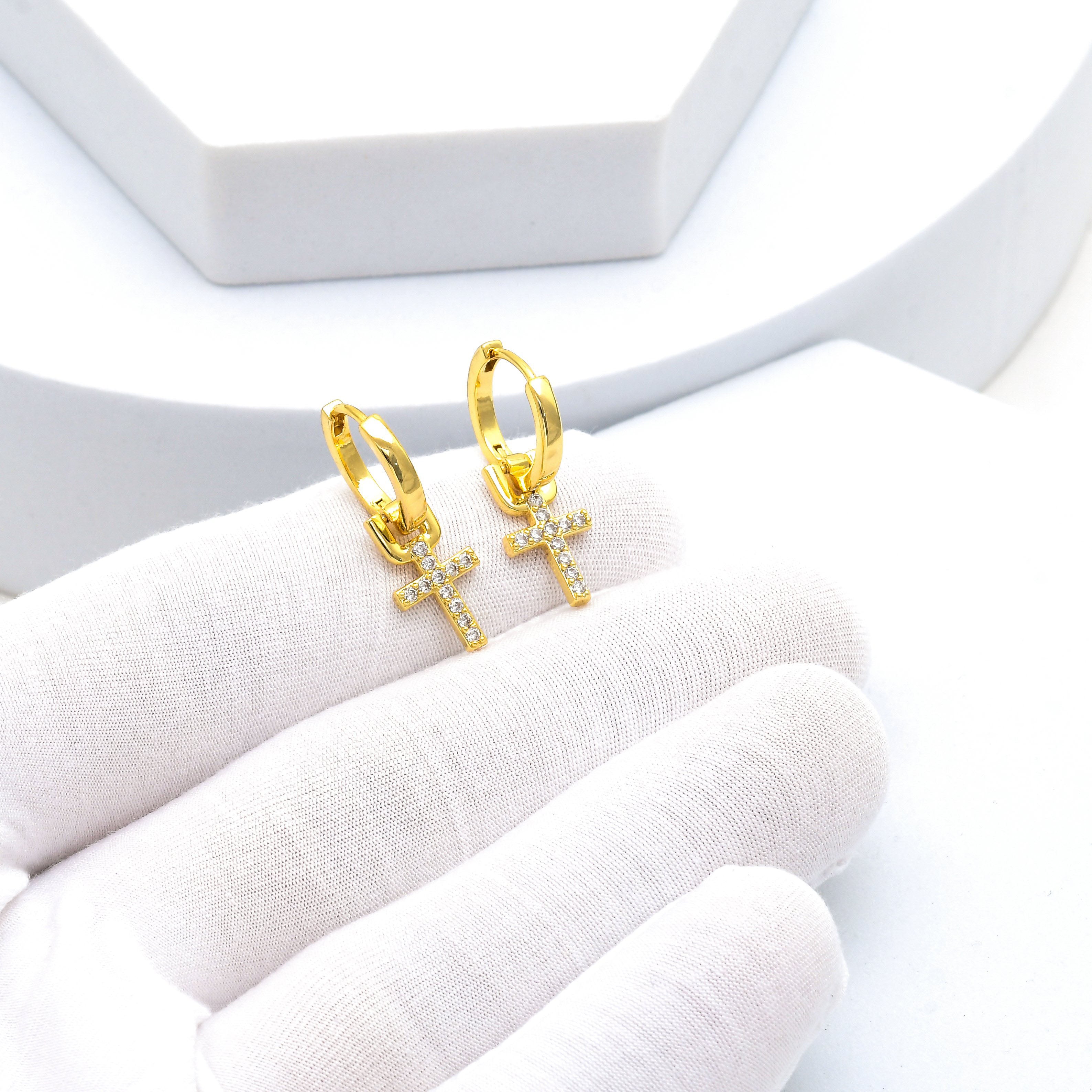 A pair of gold huggie hoop earrings with small cross charms set with clear stones, displayed on a white gloved hand by Civaro