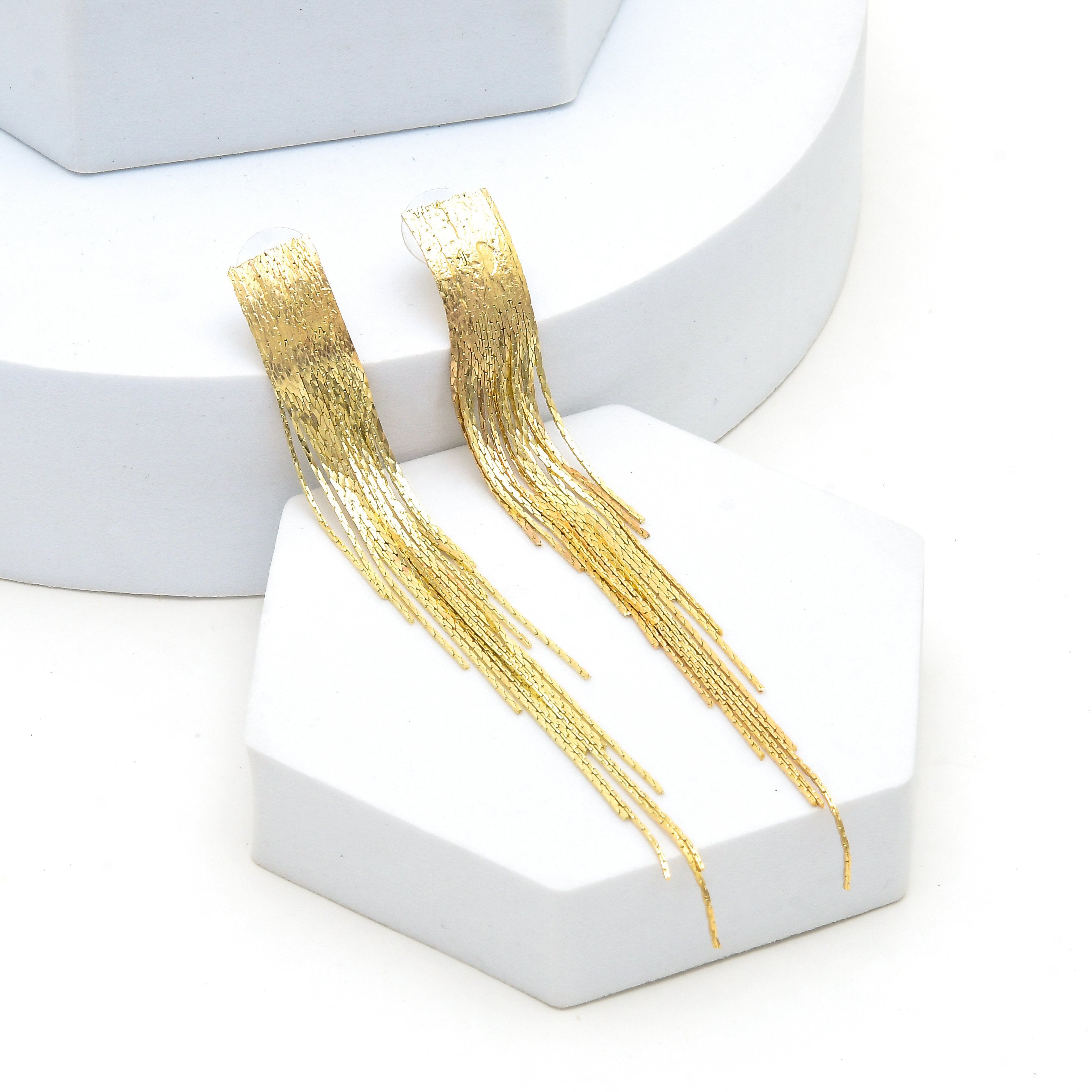 A pair of gold dangling fringe earrings displayed on white geometric stands against a clean white background by Civaro