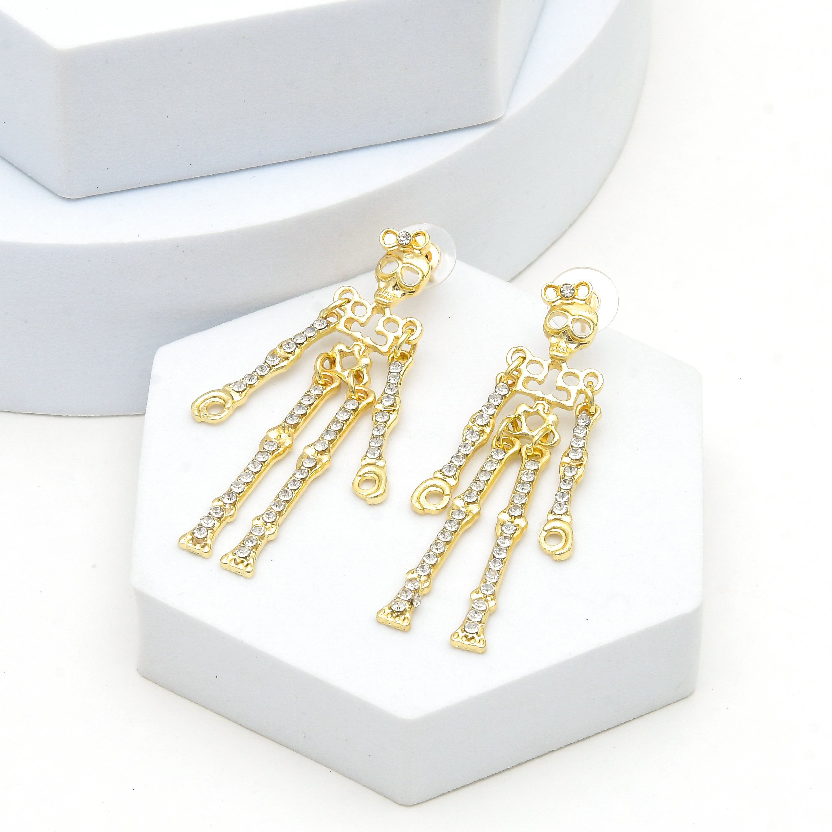 A pair of gold skeleton drop earrings with clear rhinestone-embellished limbs, displayed on a white hexagonal stand by Civaro