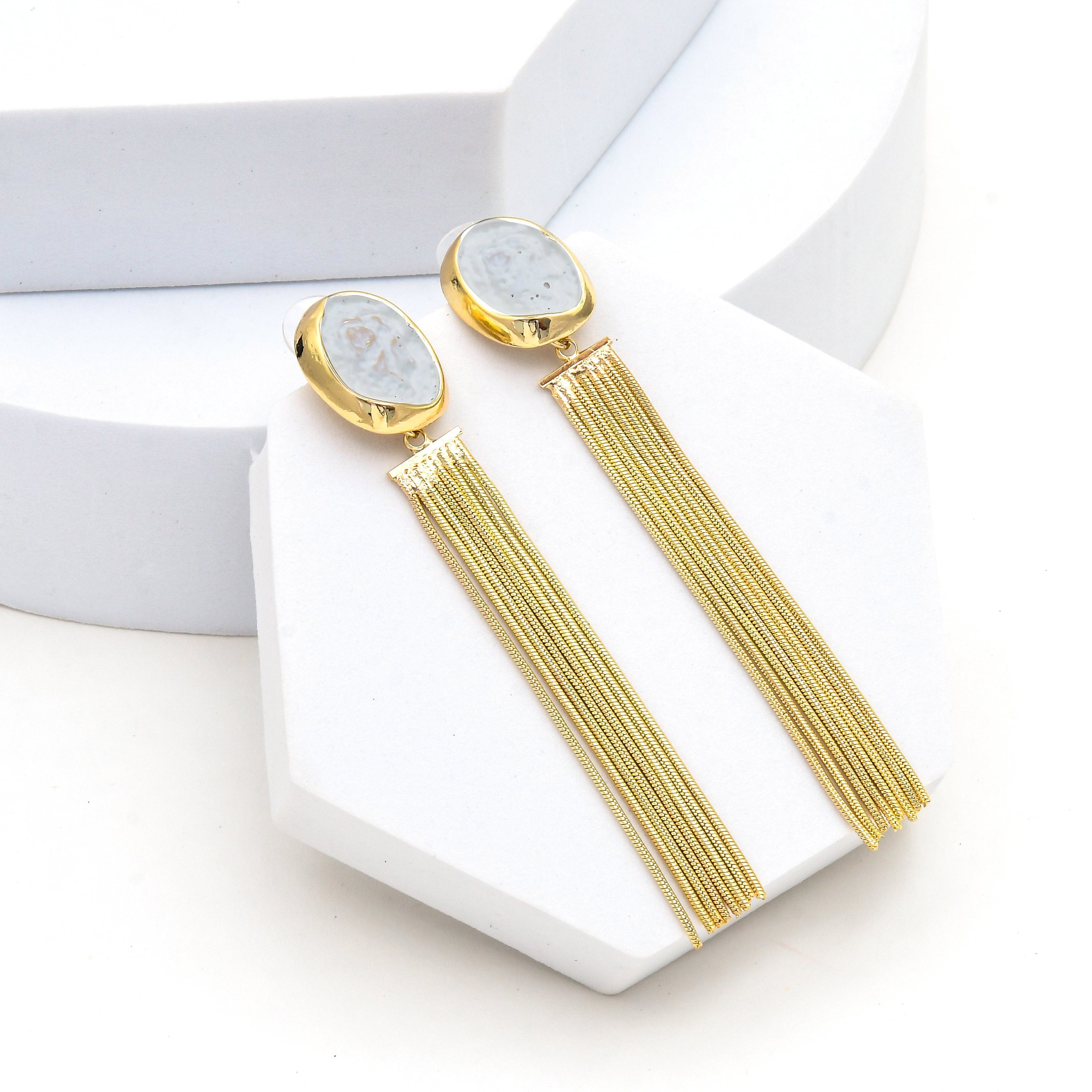 A pair of gold-toned dangle earrings with oval light blue-gray stone tops and long thin gold fringe, displayed on white geometric jewelry stands Long gold chain tassel earrings featuring an organic white marbled resin stud, elegant brass dangle earrings by Civaro