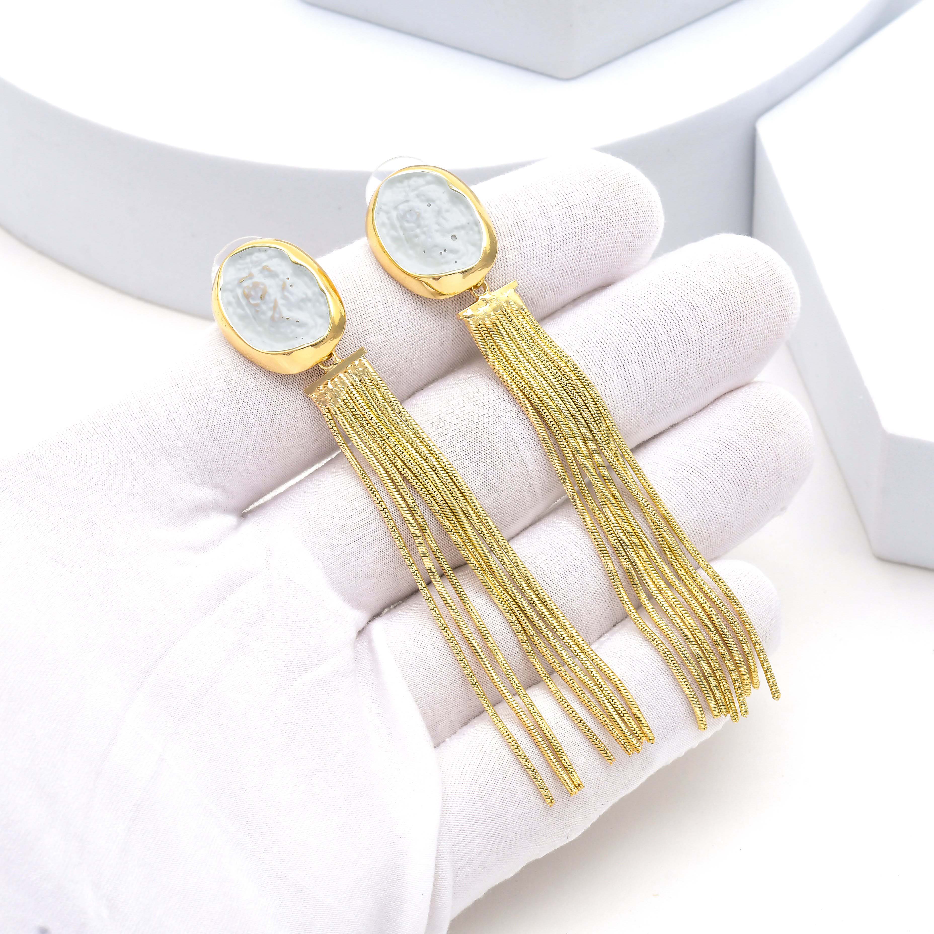 A pair of gold-toned statement dangle earrings with oval cabochon stones, displayed on a white gloved hand against a clean white background Front view of gold tassel earrings with irregular white marbled resin top and flowing chain fringe design by Civaro