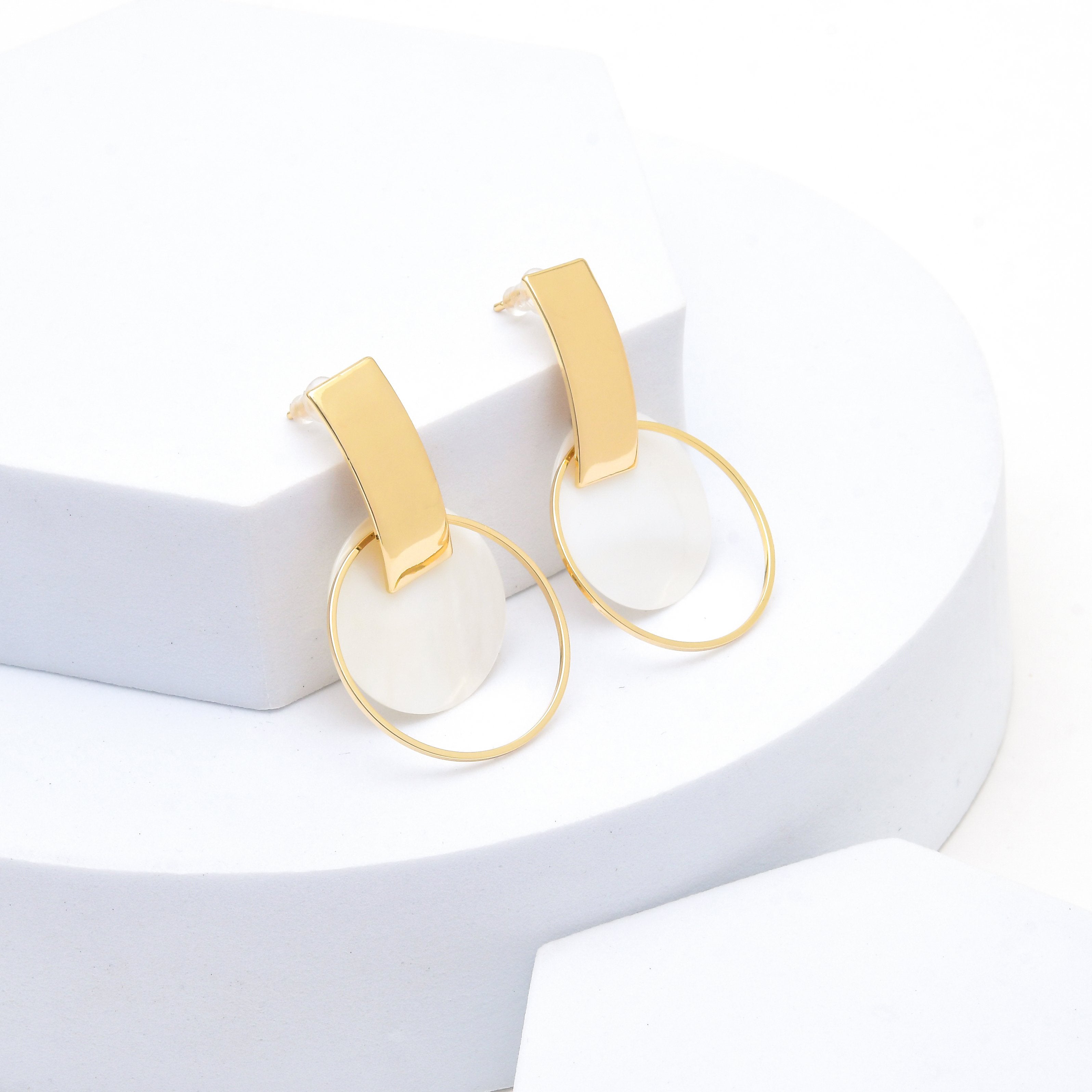 A pair of gold and white geometric dangle earrings displayed on a white tiered pedestal Premium brass gold geometric drop earrings featuring a natural shell centerpiece with a polished gold tone finish by Civaro