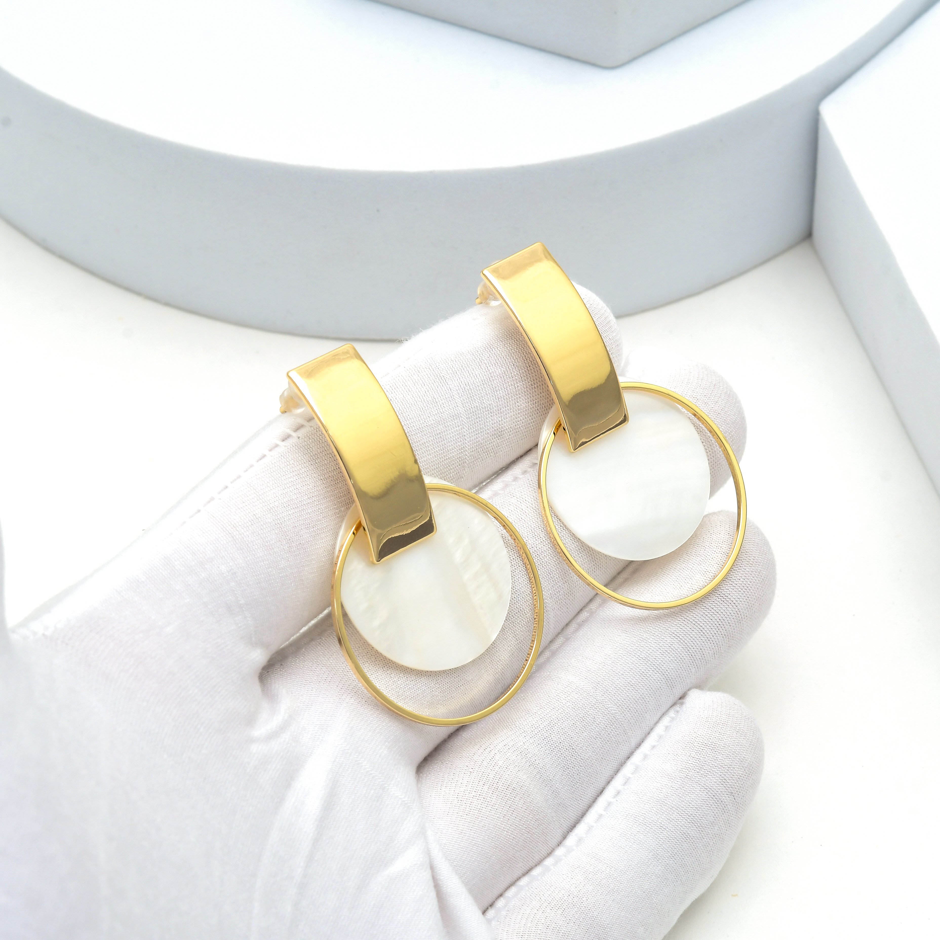 A pair of gold hoop earrings with white circular disc drops, held in a white gloved hand against a clean white background. Modern lightweight brass dangle earrings featuring luminous shell detail and minimalist gold design by Civaro