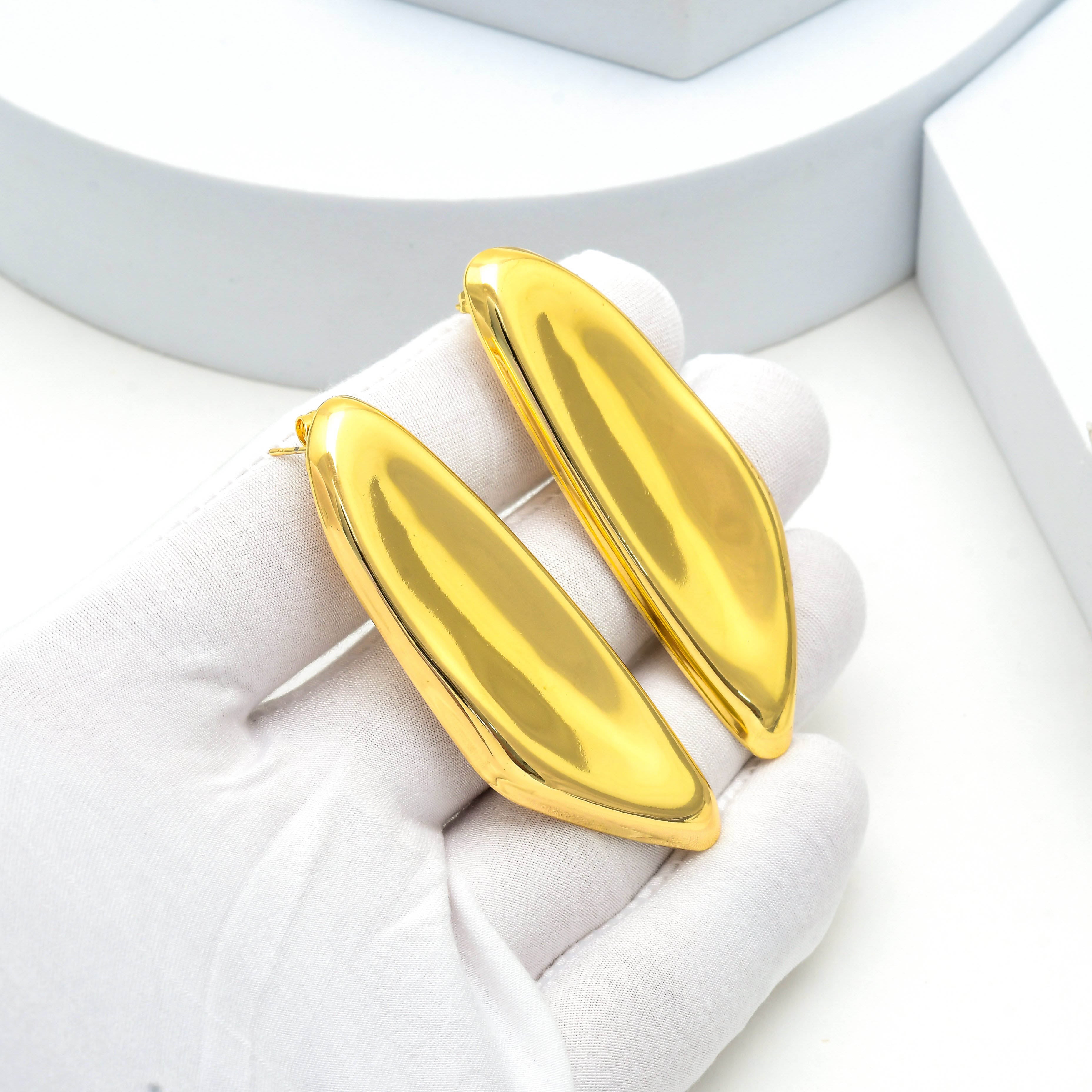 A pair of large, polished gold leaf-shaped earrings held in a white gloved hand against a clean white background Close up of polished stainless steel statement earrings showcasing the smooth sculptural surface and secure push back stud design by Civaro