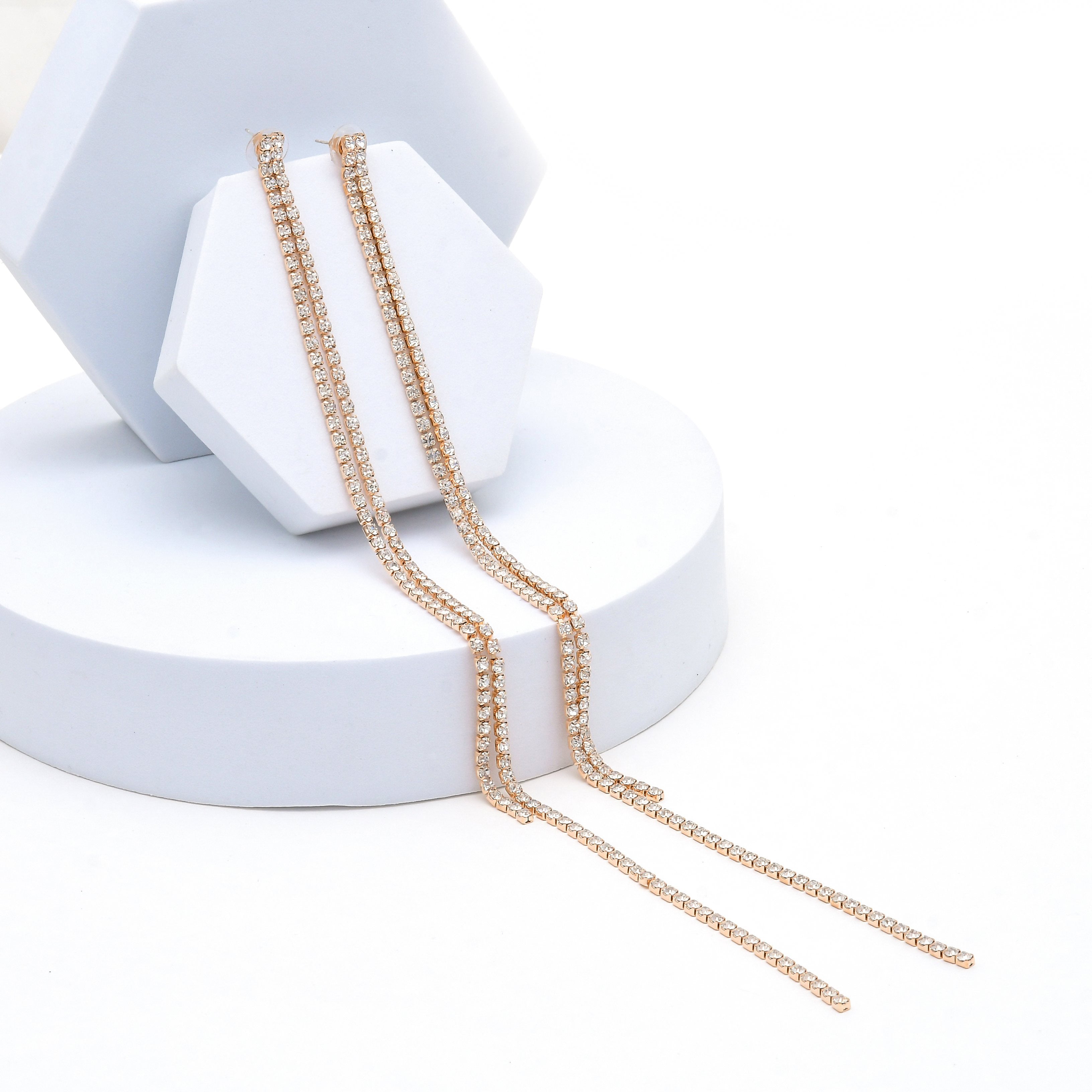 A pair of delicate gold-toned chain earring  draped over a white geometric display stand on a clean white background. Luxury-inspired tassel earrings with an 18K rose gold tone finish and elegant linear crystal design by Civaro