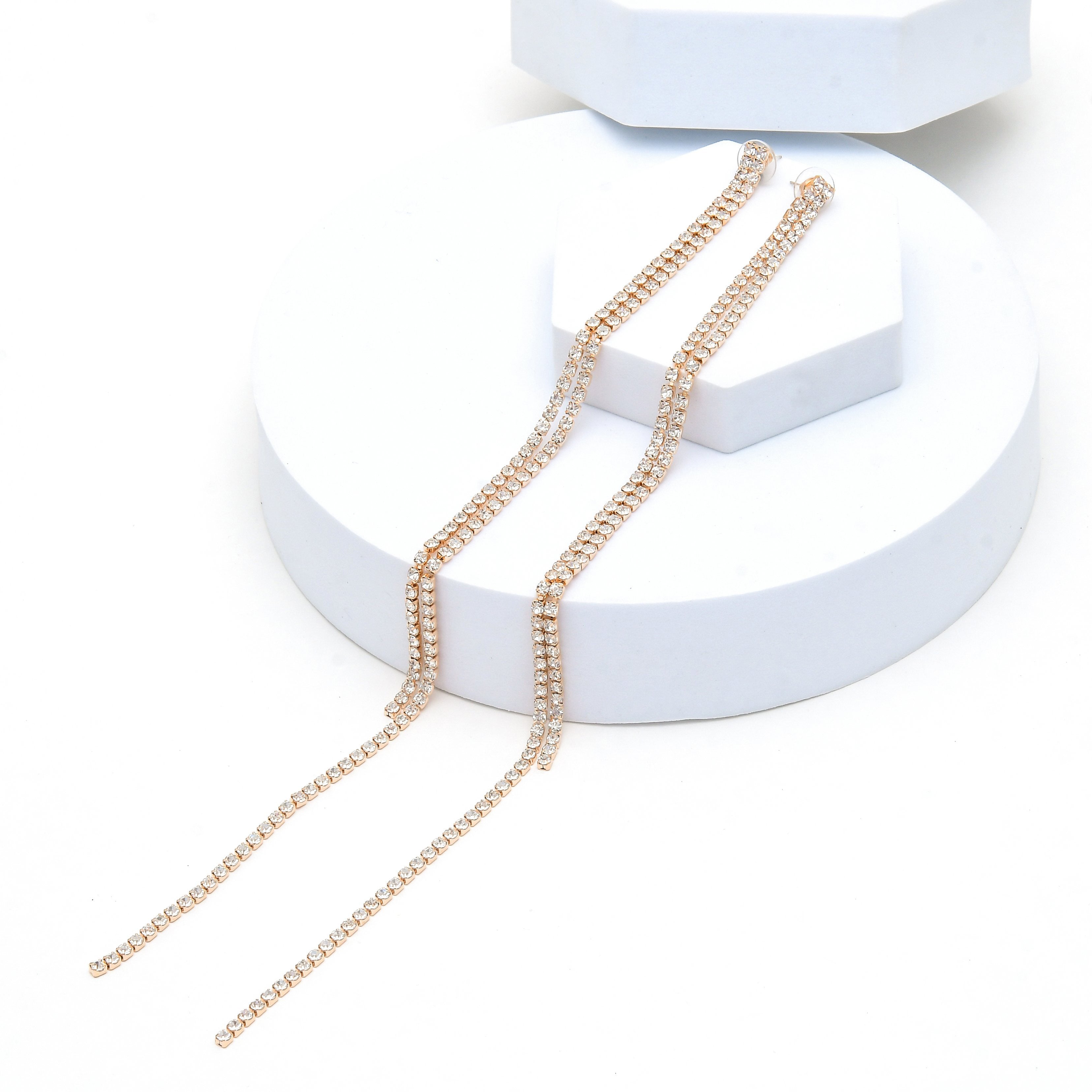 A pair of long, delicate gold chain necklaces embellished with small clear rhinestones, draped over a white geometric display stand on a white background. Rose gold tassel earrings designed for weddings, anniversaries, formal occasions and sophisticated everyday wear by Civaro
