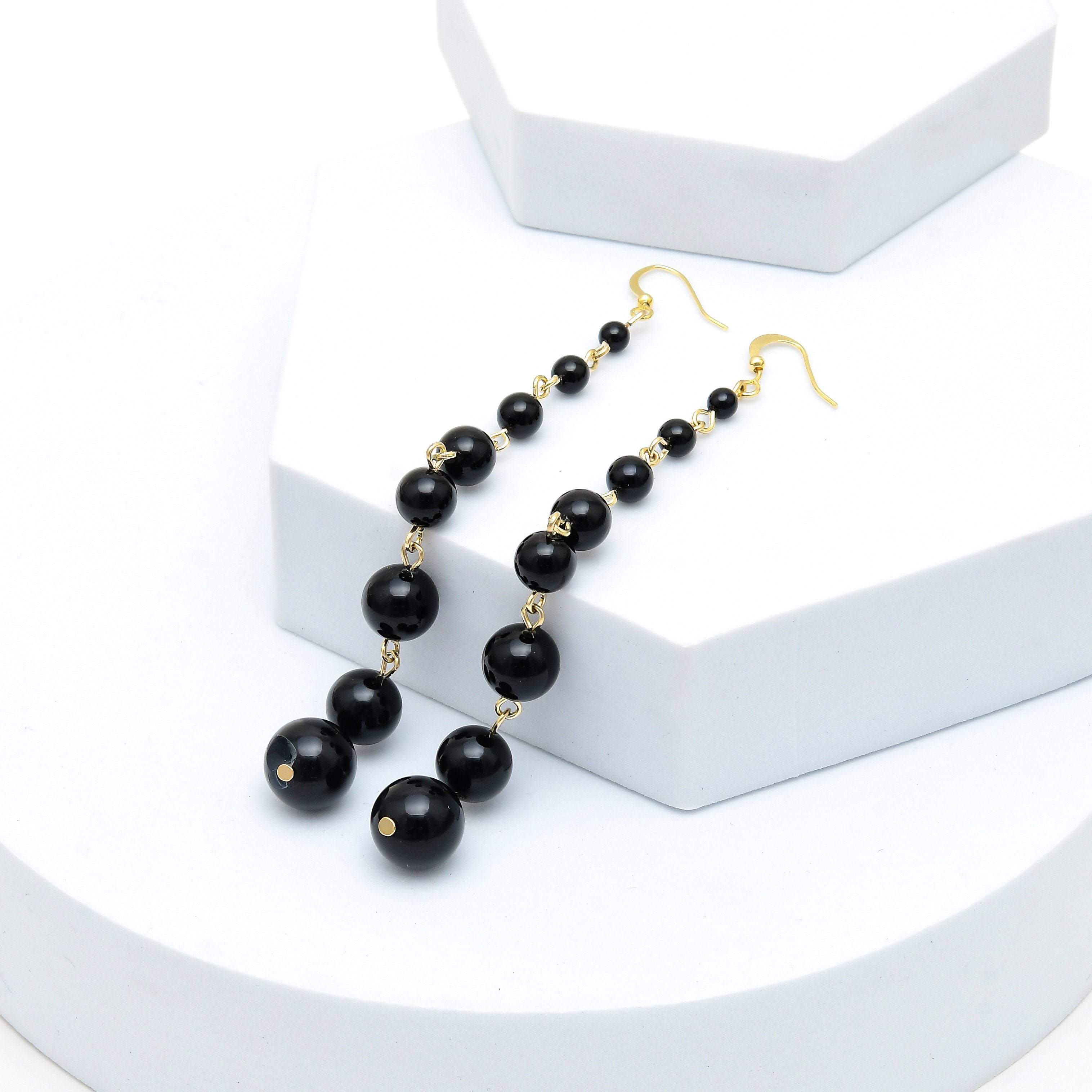 A pair of long drop earrings with glossy black round beads and small gold spacer rings, hanging from gold fishhook ear wires, displayed on a white geometric riser, Elegant black pearl dangle earrings showcasing glossy graduated pearls connected by polished gold tone stainless steel links by Civaro