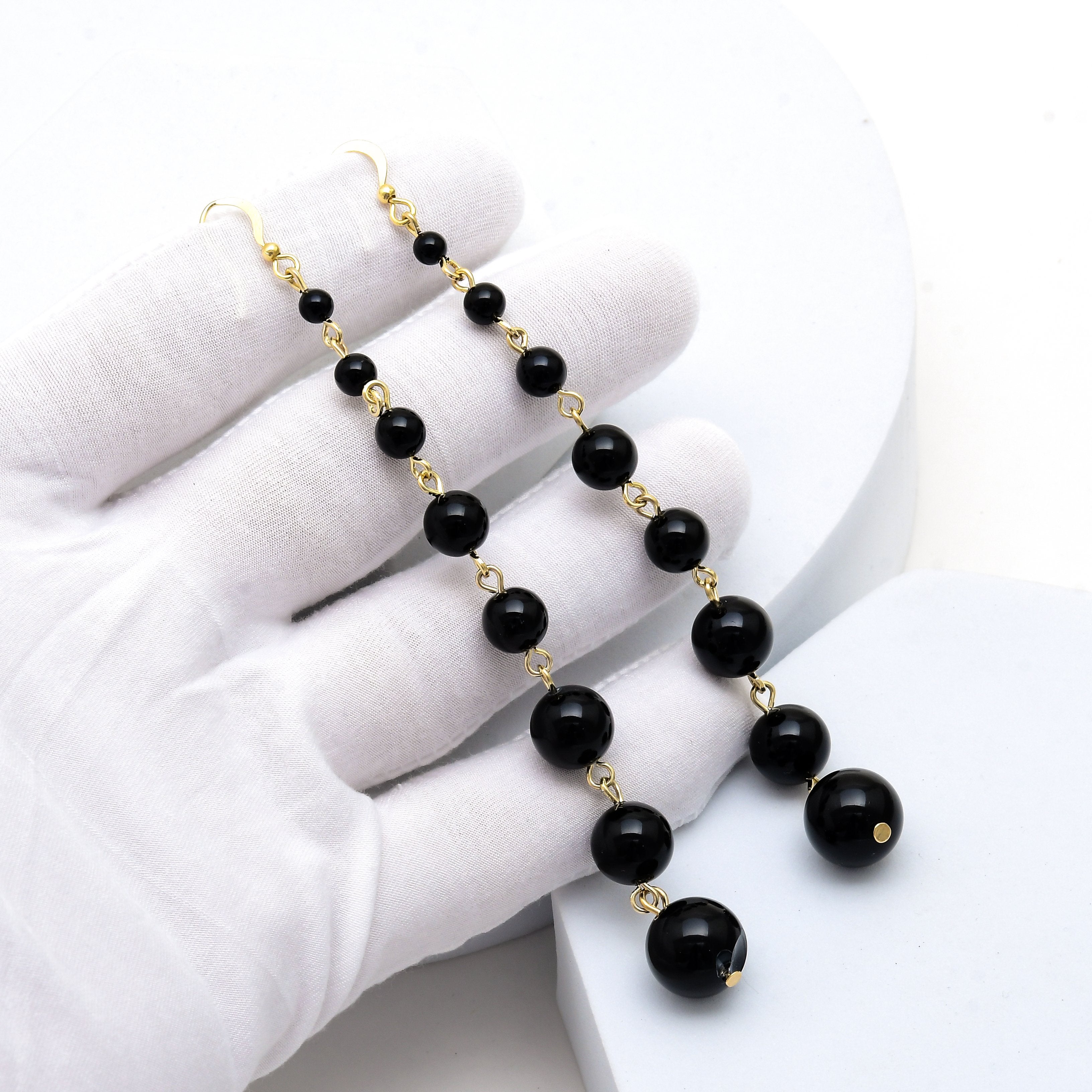 A pair of long black beaded drop earrings with gold-tone links and hooks, displayed on a white gloved hand against a clean white background, Lightweight black pearl statement earrings with cascading bead detailing and durable stainless steel construction in a gold tone finish by Civaro