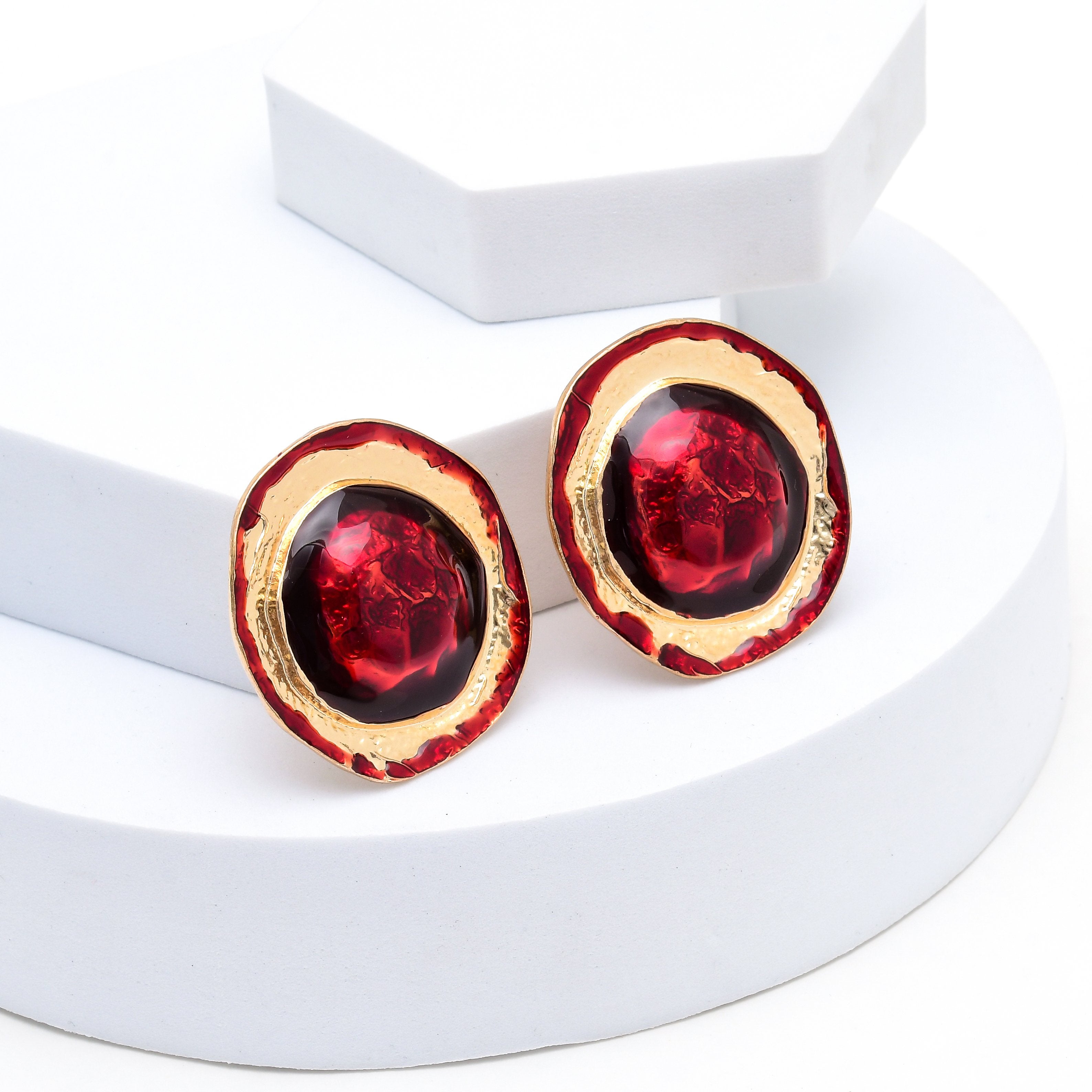 A pair of round stud earrings with textured gold-toned edges, deep red outer rings, and glossy black centers featuring raised red details, displayed on a white geometric stand. Premium alloy and brass clip-on earrings featuring bold red accents and textured gold-tone detailing by Civaro.
