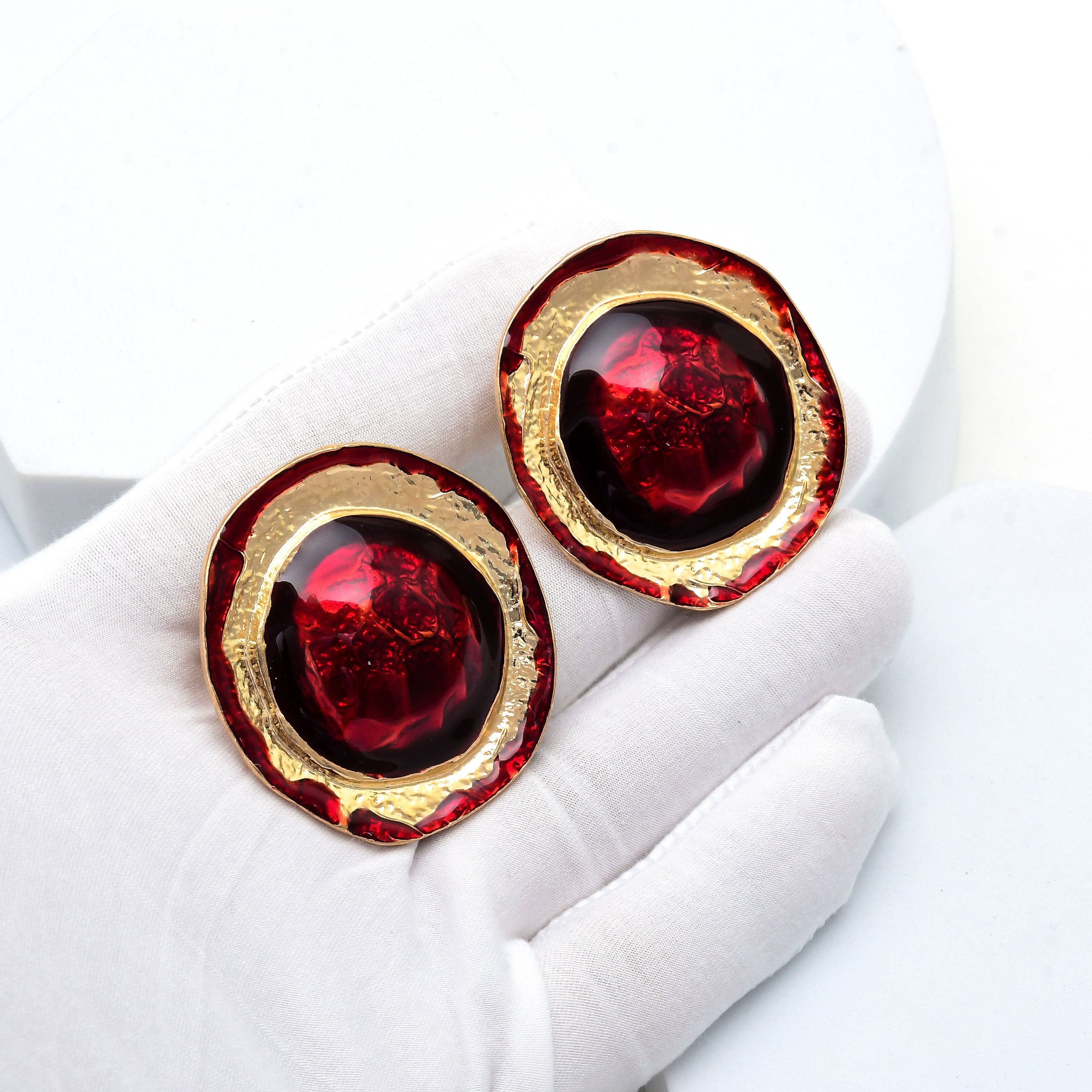 A pair of round gold-tone earrings with deep red marbled centers, displayed on a white gloved hand against a white background. Oversized red clip earrings designed for party, wedding and evening occasion styling by Civaro.
