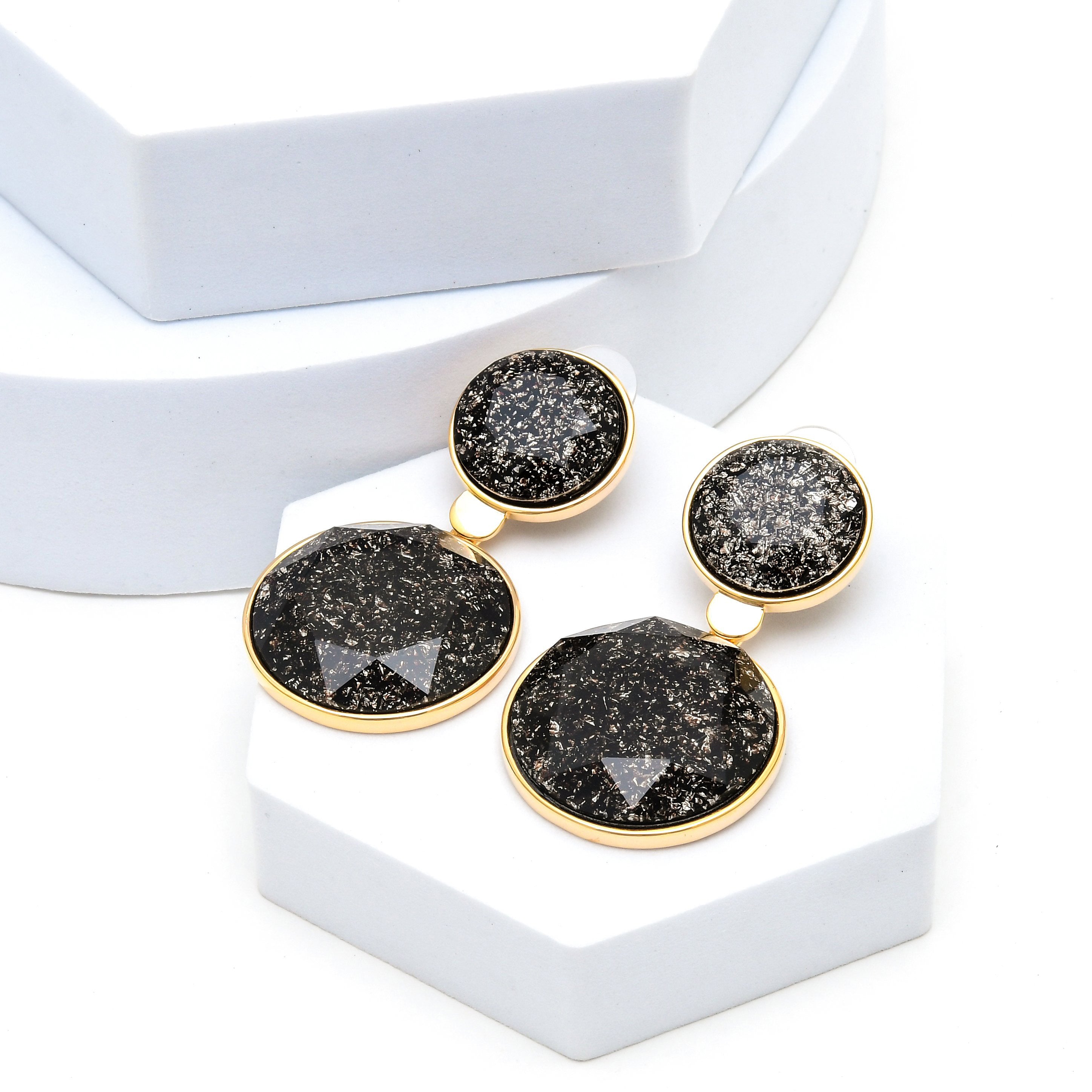 Modern black statement earrings designed with faceted decorative centres and premium alloy construction by Civaro.
 A pair of gold-tone drop earrings with round black druzy stone pendants, displayed on white geometric risers against a white background.