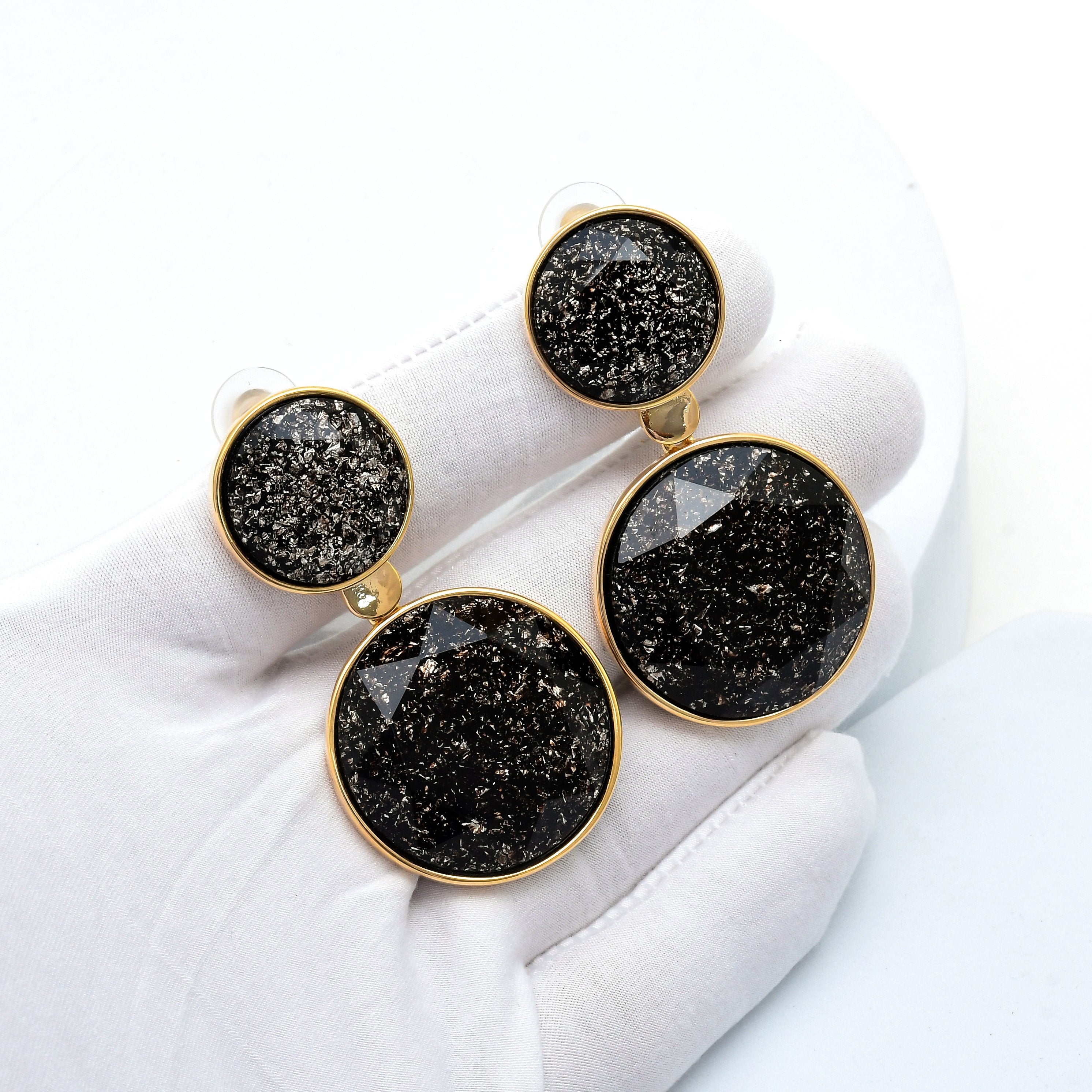 A pair of round drop earrings with black druzy stones and gold-tone bezels, displayed on a white gloved hand against a white background. Premium alloy and brass black statement earrings featuring faceted decorative accents and polished gold tone borders by Civaro.
