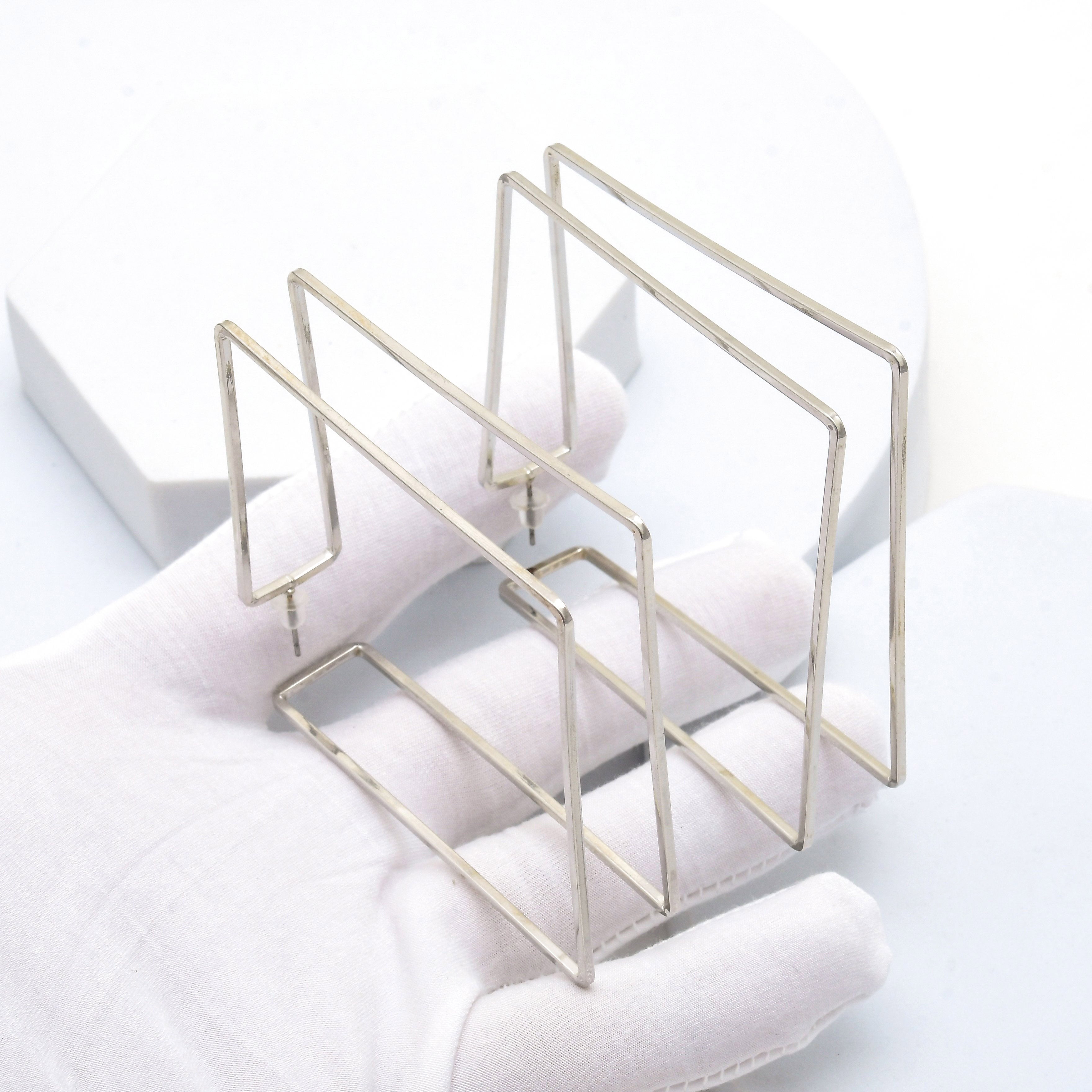 Women's oversized geometric silver earrings crafted from polished stainless steel with a lightweight design by Civaro.
 A pair of silver-toned metal wire jewelry display stands held in a white gloved hand against a clean white background.