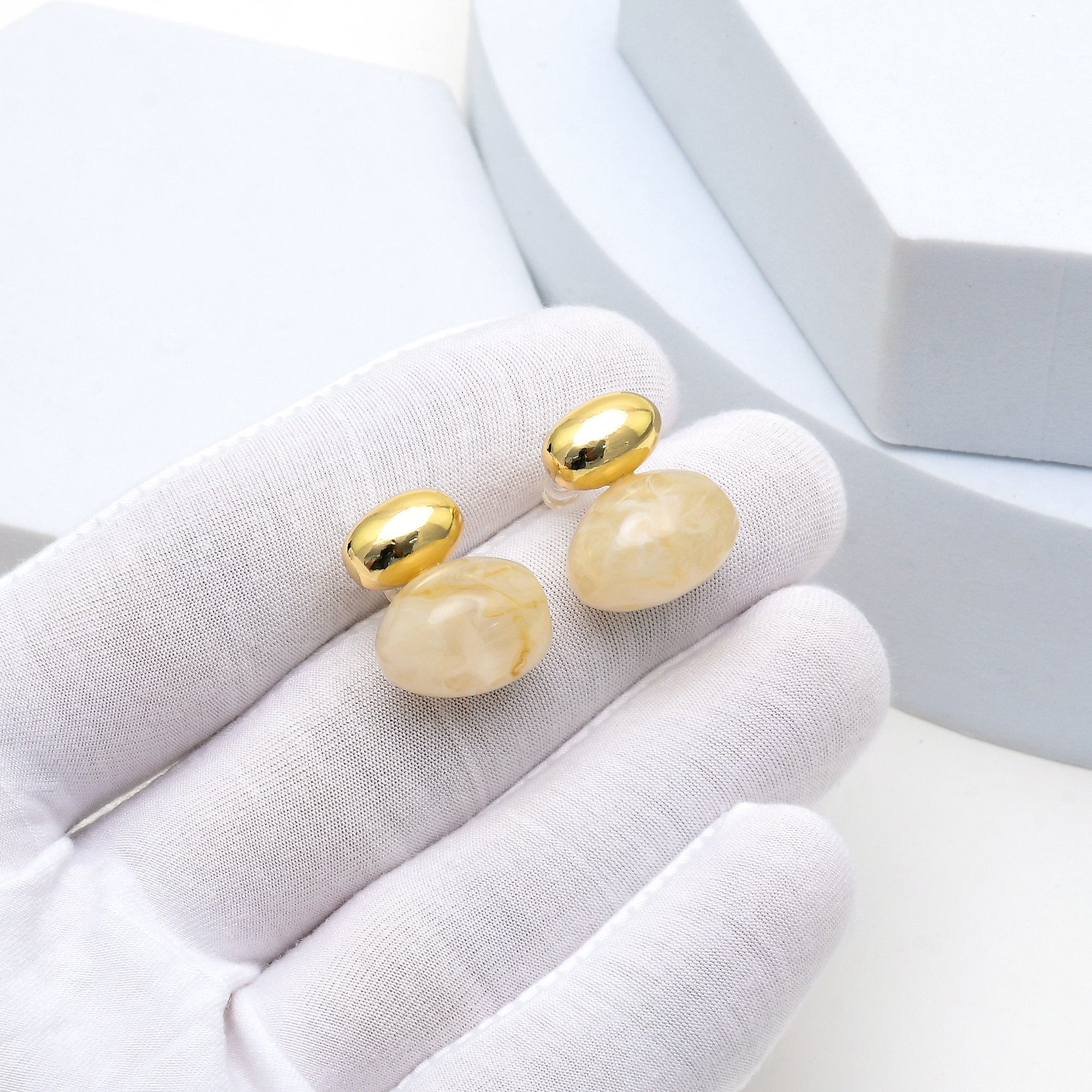 A pair of gold stud earrings with oval cream-colored stone drops, held on a white gloved hand against a light gray jewelry display background by Civaro