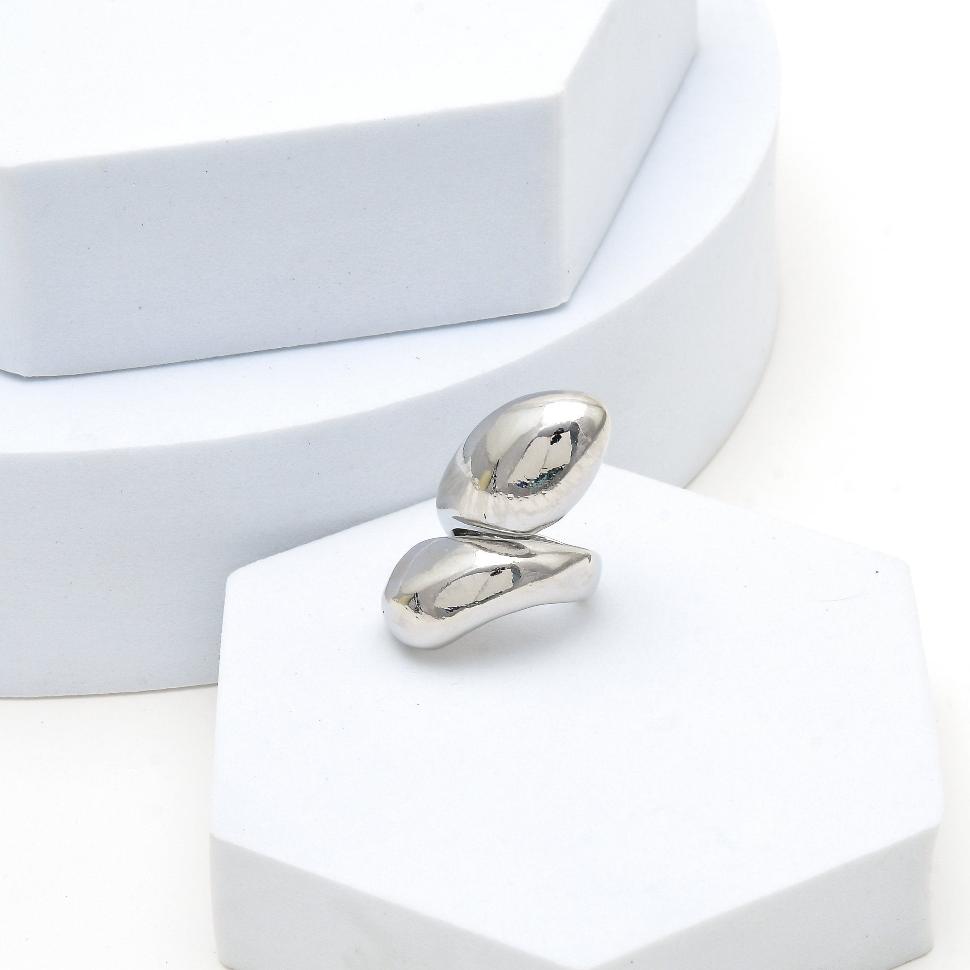 A polished silver abstract ring with two curved, organic shapes, displayed on a white hexagonal pedestal.