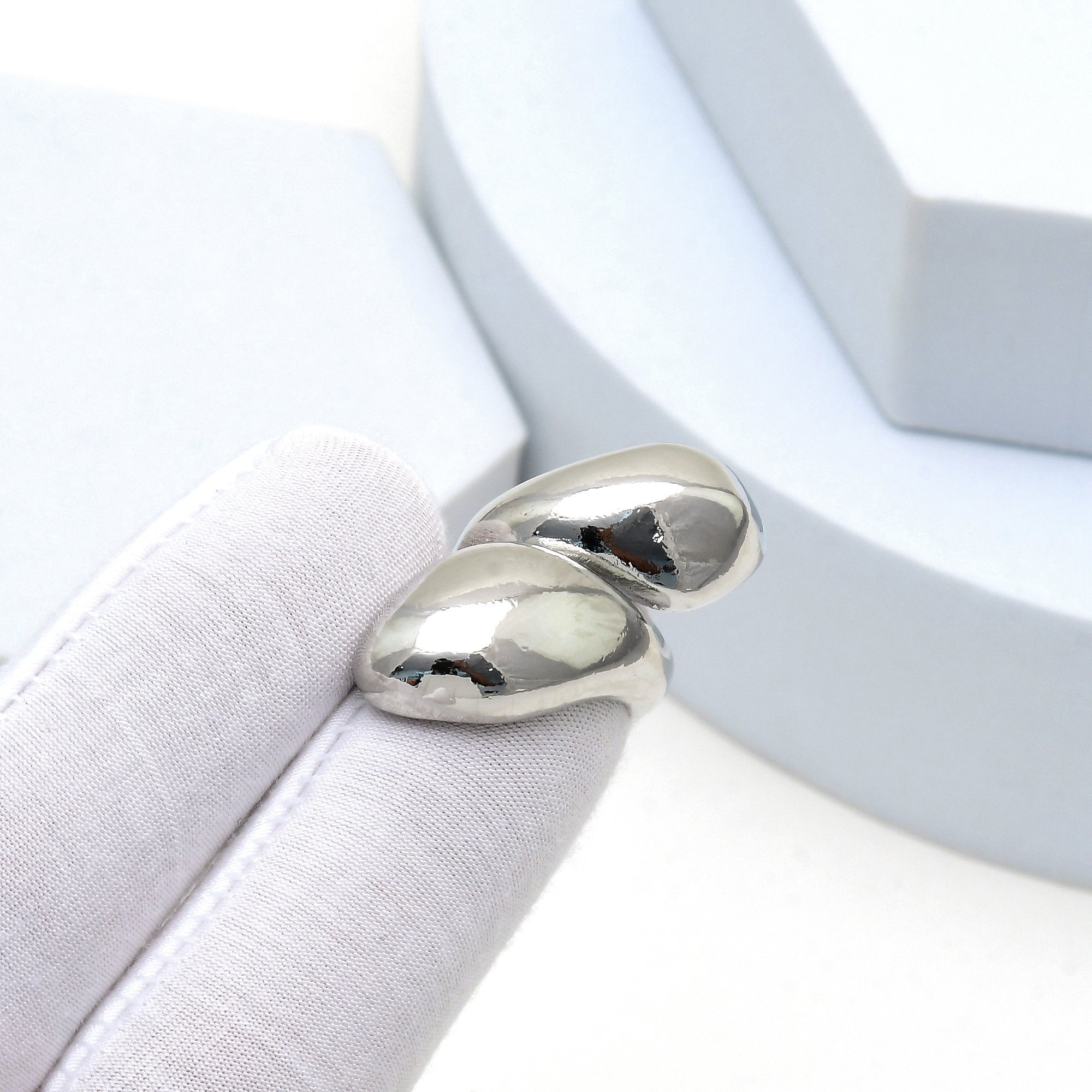 A close-up of a polished silver ring with a double-oval design, held on a white fabric-covered display stand.