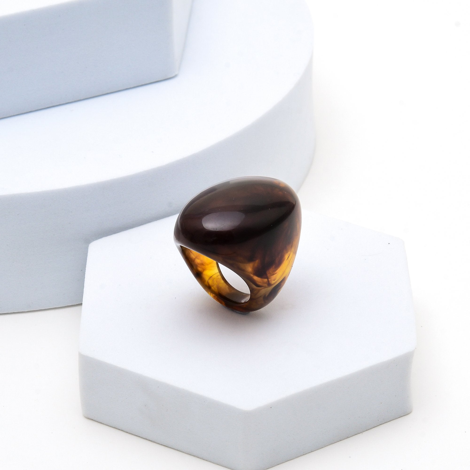 A chunky organic-shaped ring made of glossy amber-brown translucent resin, displayed on a light gray geometric pedestal against a white background.