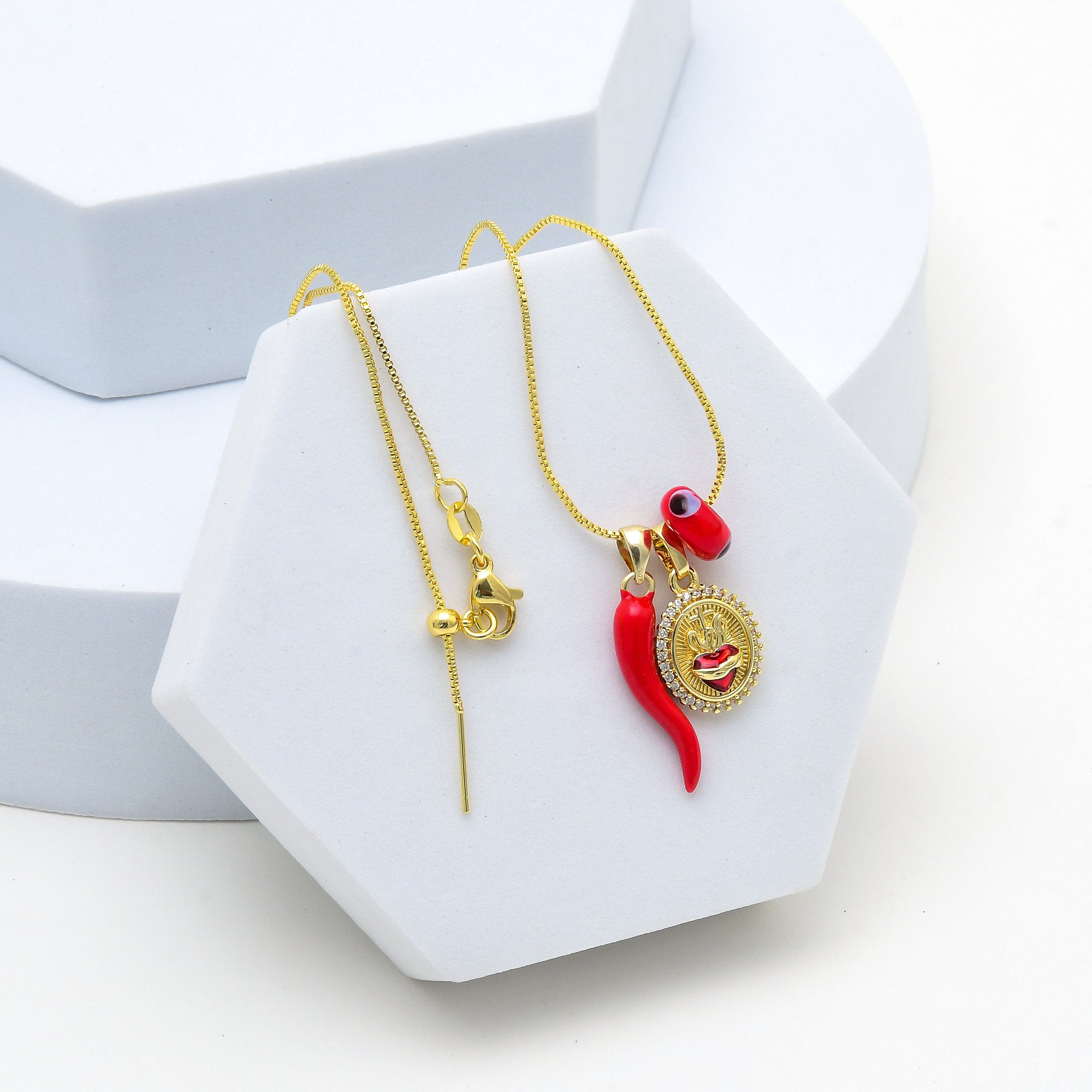 Gold-tone necklace and matching drop earring with a red chili pepper charm, a red evil eye bead, and a round gold medallion pendant, displayed on a white hexagonal stand. Gold symbolic pendant necklace with sacred heart and evil eye bead by Civaro