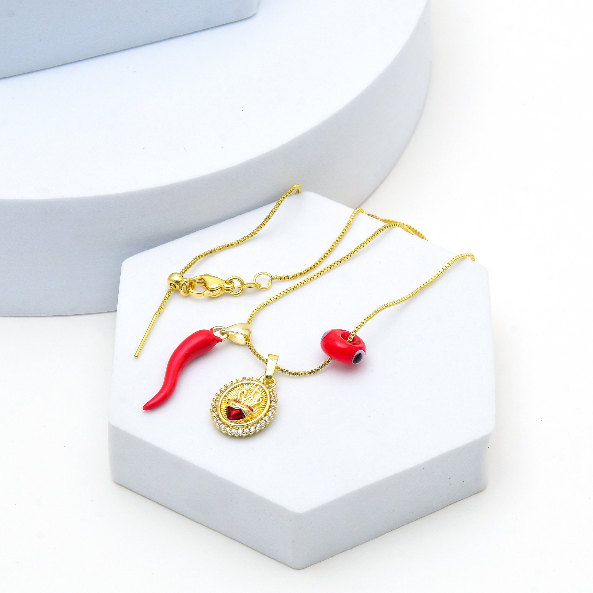 Layering necklace featuring Sacred Heart and chili pendant
 A gold chain necklace with two red chili pepper charms, displayed on a white hexagonal stand by Civaro