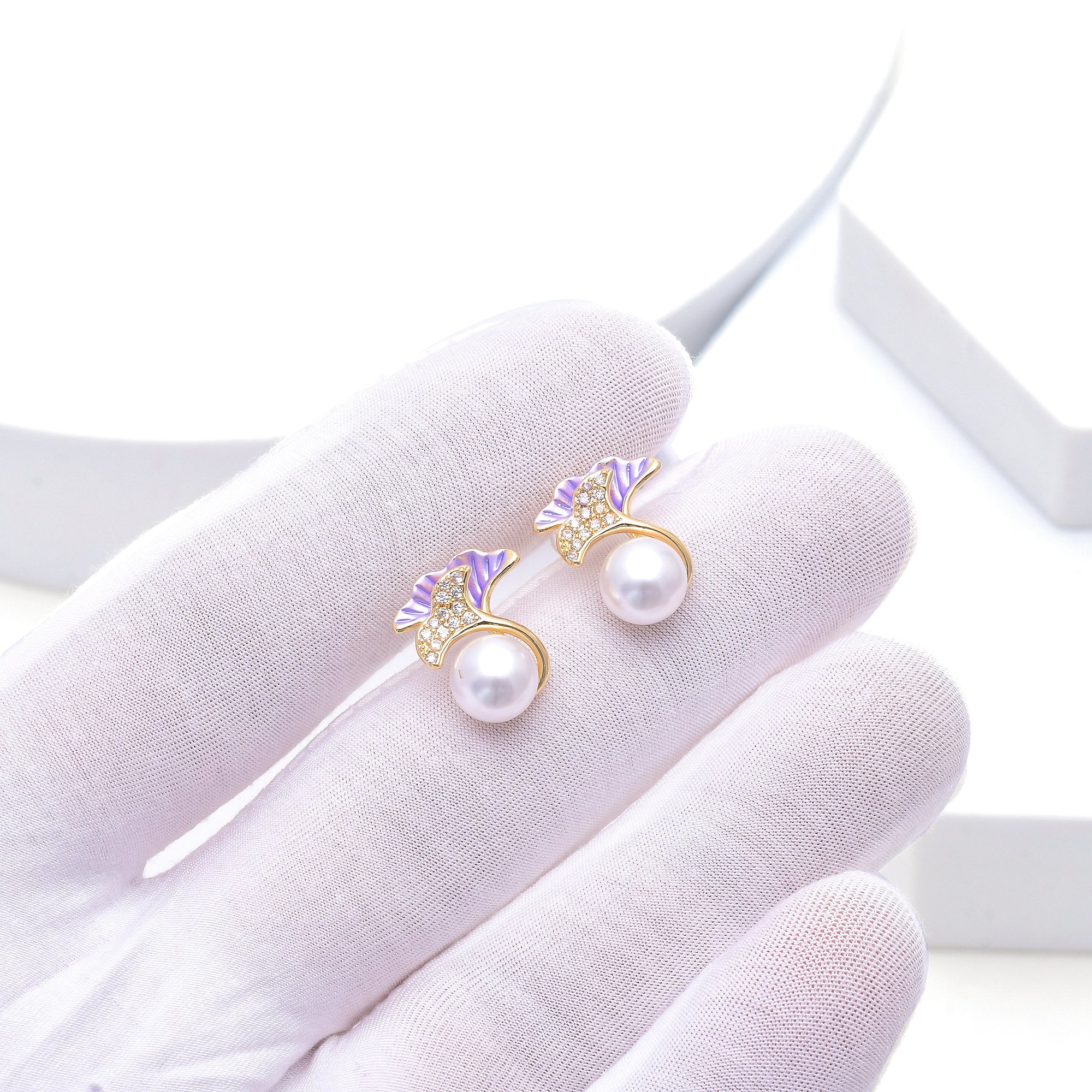 Petite floral earrings featuring soft lilac petals, clear decorative accents and pearl-style centres by Civaro.
A pair of gold-tone earrings with white pearl drops and purple enamel leaf details, held on a white gloved finger.