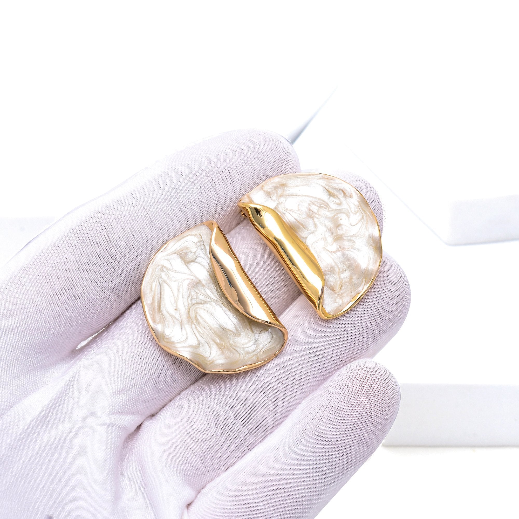Sculptural cream earrings featuring organic curved shapes and polished gold-tone detailing by Civaro.
A pair of gold-tone earrings with a marbled cream resin inlay, held in a white-gloved hand against a bright white background.