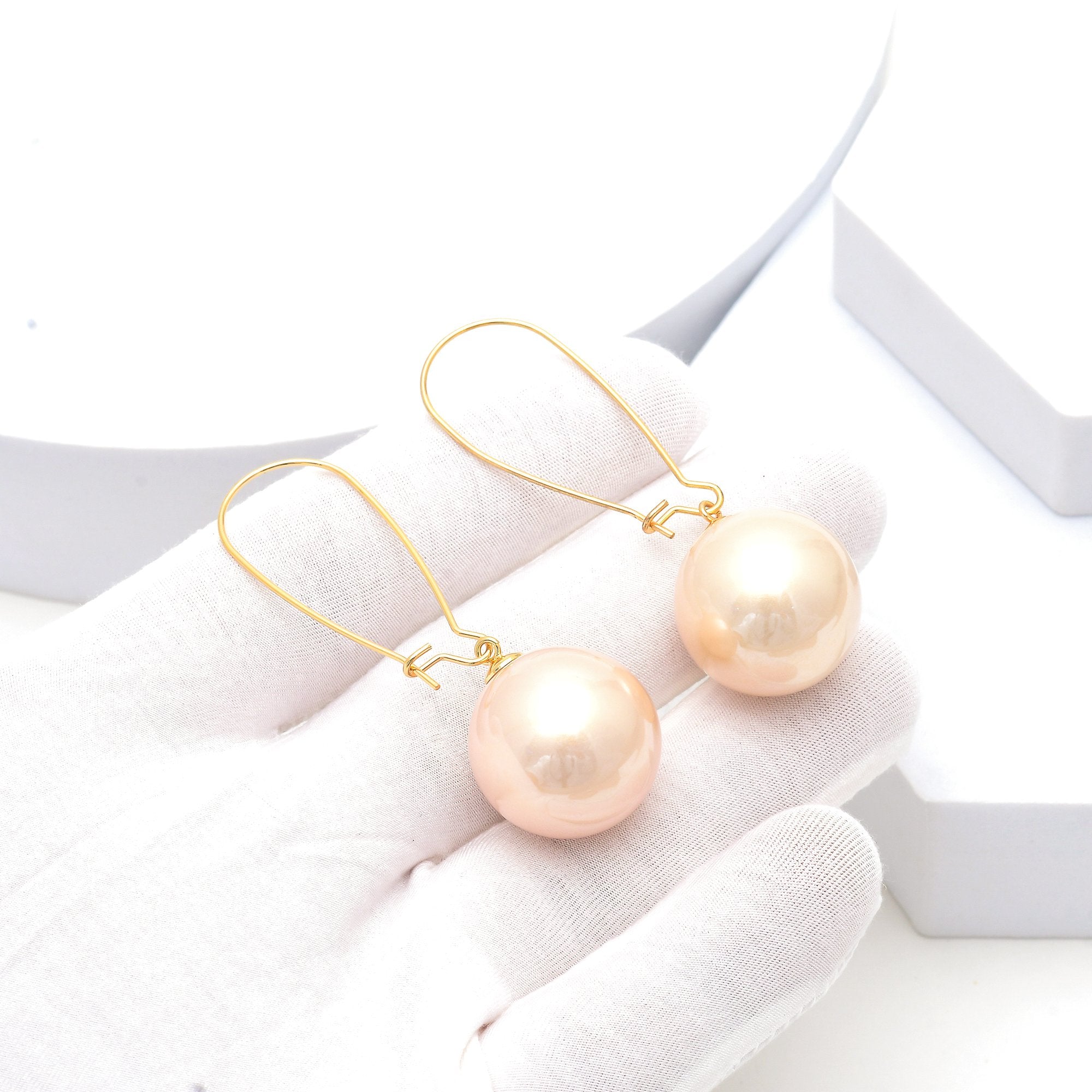 Elegant pearl-style earrings for women featuring large luminous round drops suspended from fine hooks by Civaro.
A pair of gold-tone drop earrings with large round peach-pink pearls, displayed on a white gloved hand against a clean white background.
