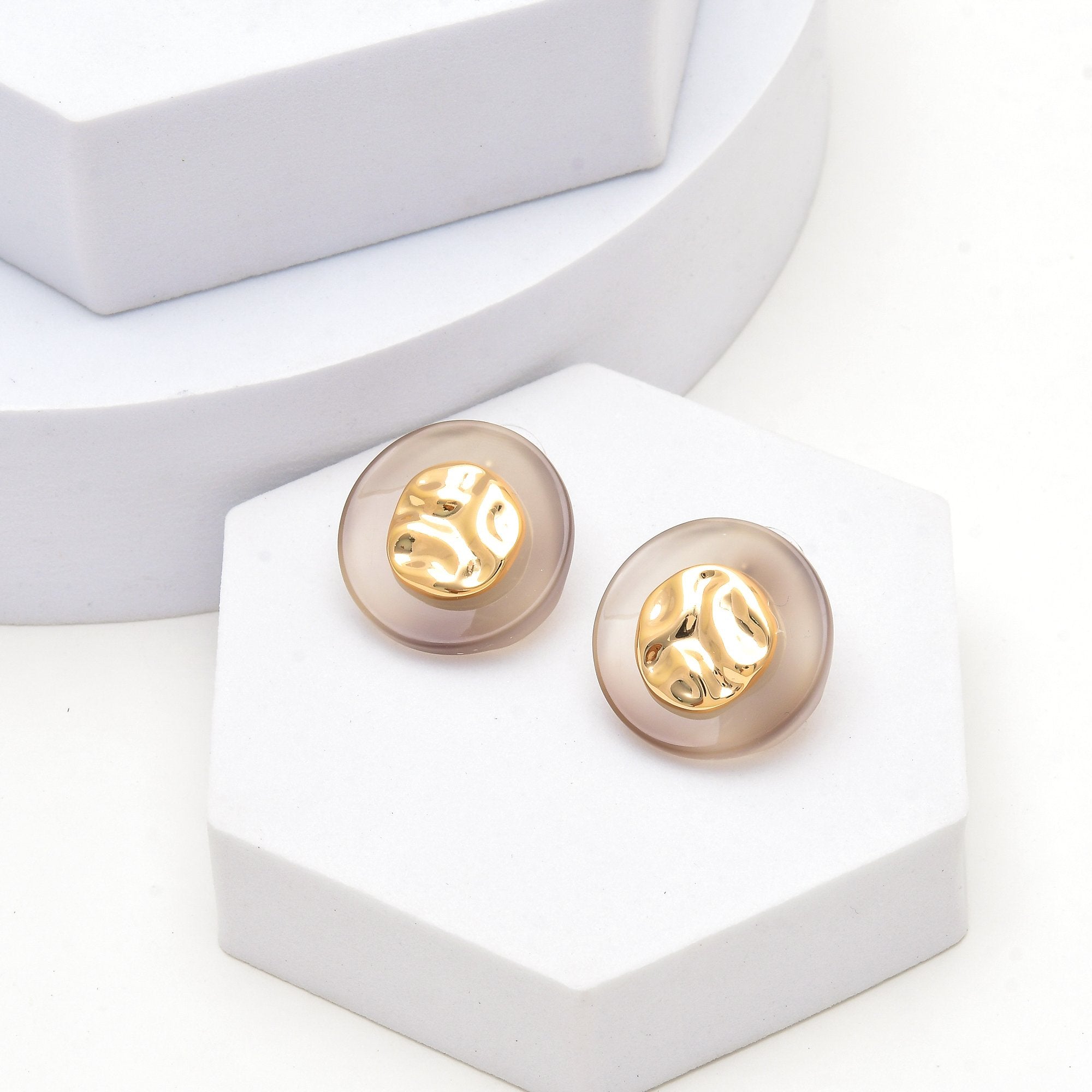 Gold round stud earrings featuring a soft taupe decorative outer section for modern everyday styling by Civaro.
A pair of round stud earrings with translucent smoky beige outer domes and gold-toned inner discs, displayed on white geometric blocks.