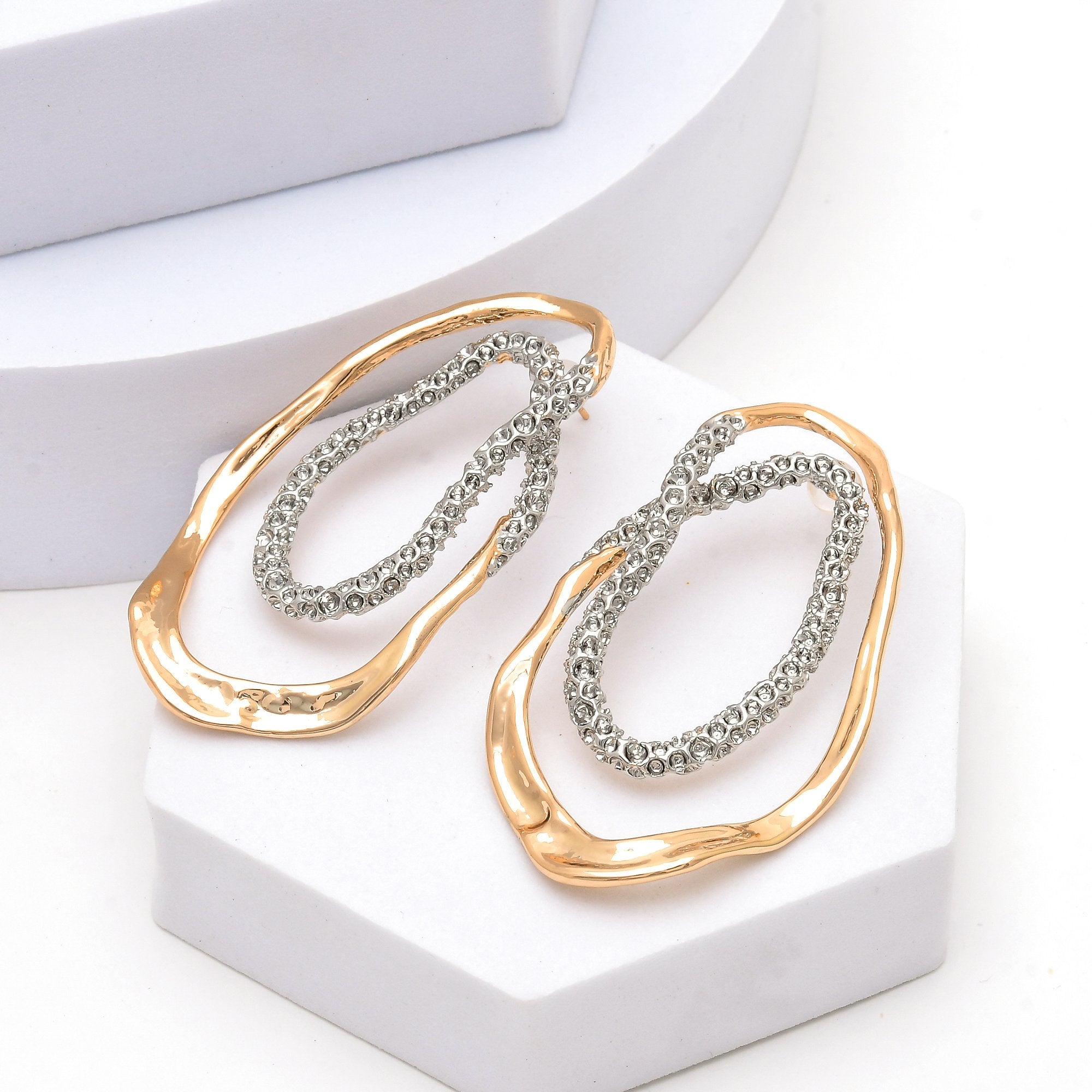 Abstract mixed metal earrings featuring sculptural gold-tone curves and detailed silver-tone accents by Civaro.
A pair of abstract statement earrings with textured gold and silver wavy borders, displayed on a white geometric stand.