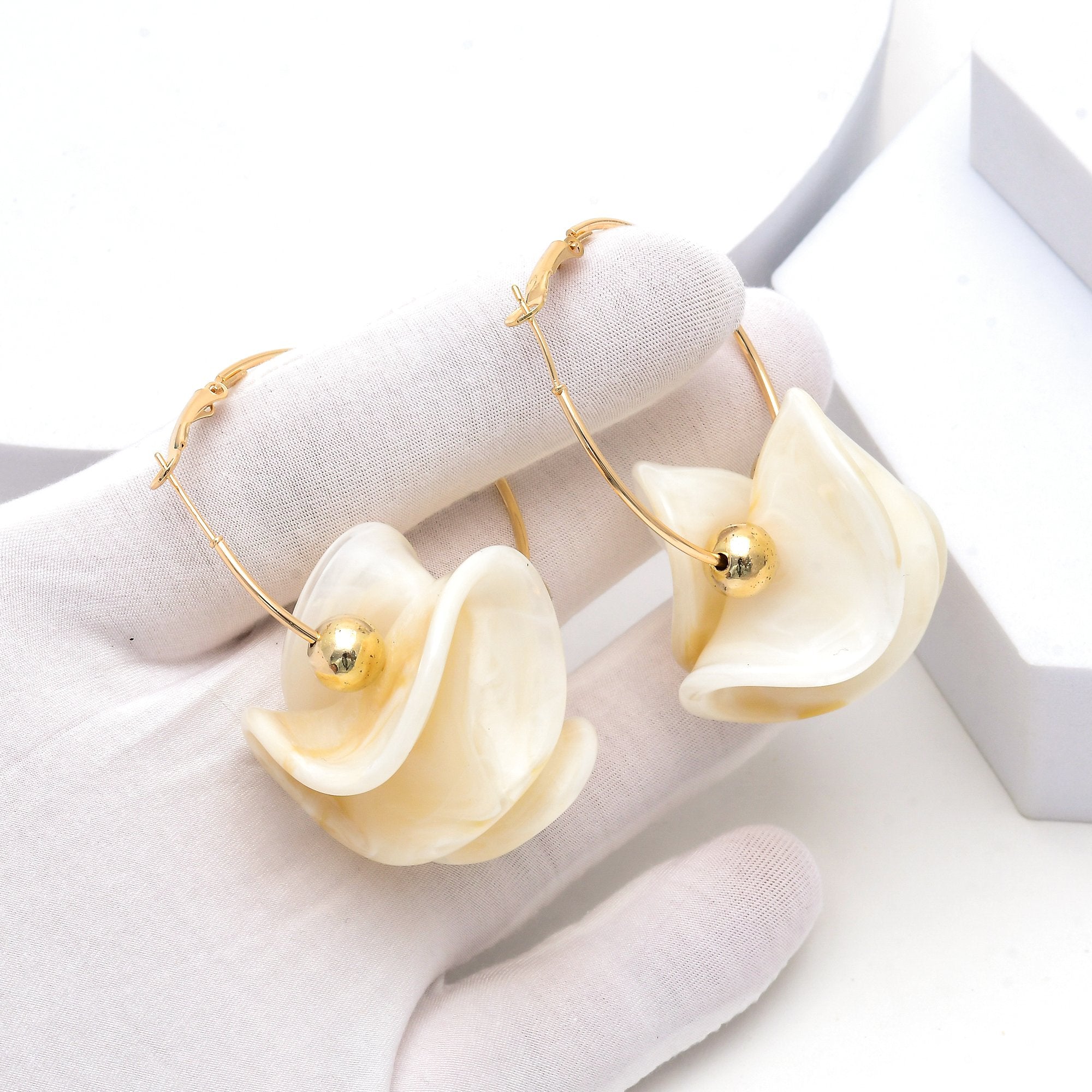 .Side view showing the layered acrylic petal design and gold-tone hoop fastening by Civaro.
A pair of gold hoop earrings with layered cream-colored shell-like flower drops, displayed on a white gloved hand.