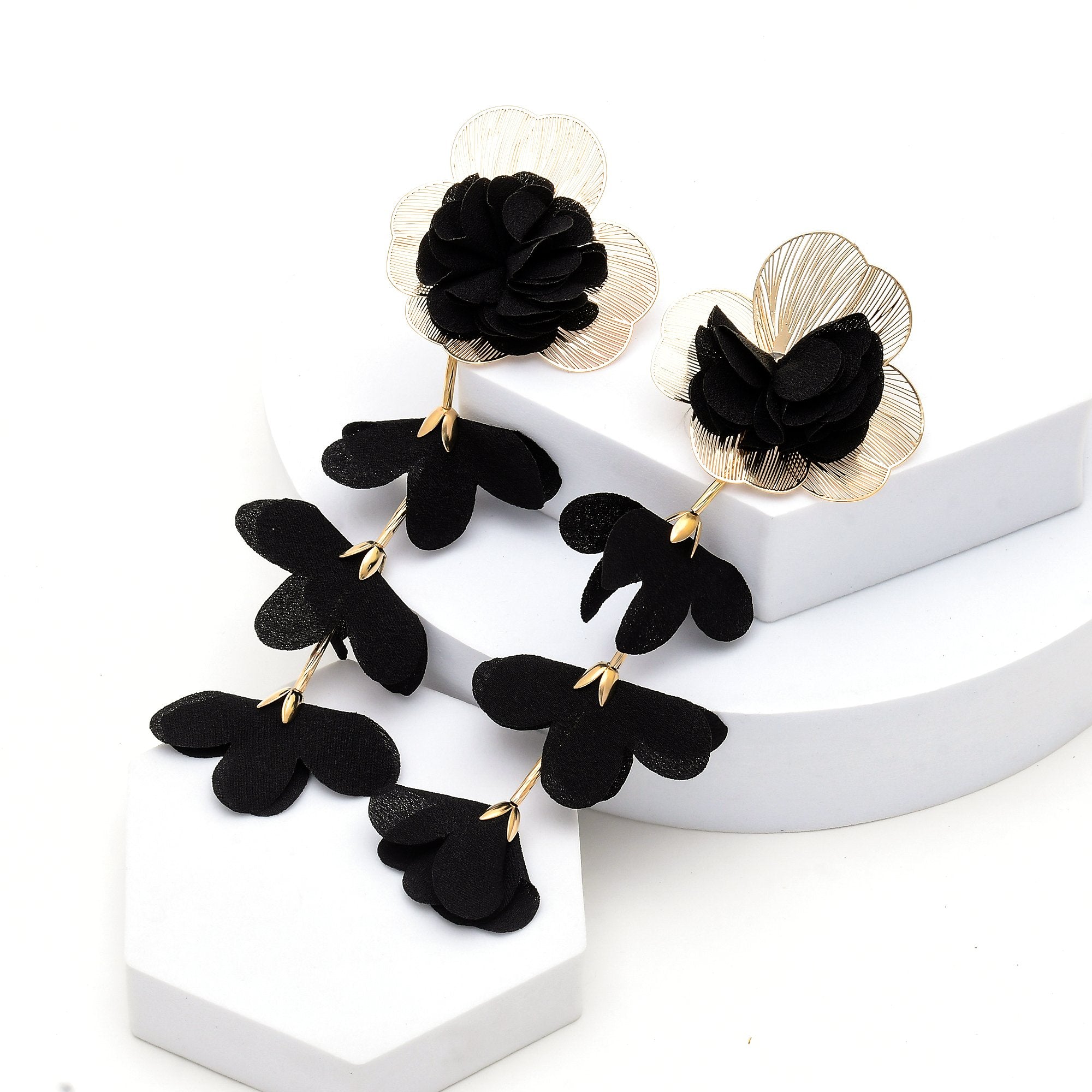 Close-up of the layered black textile flower set inside a delicate gold-tone floral frame by Civaro.Black and gold flower drop earrings displayed on white geometric risers against a white background.
