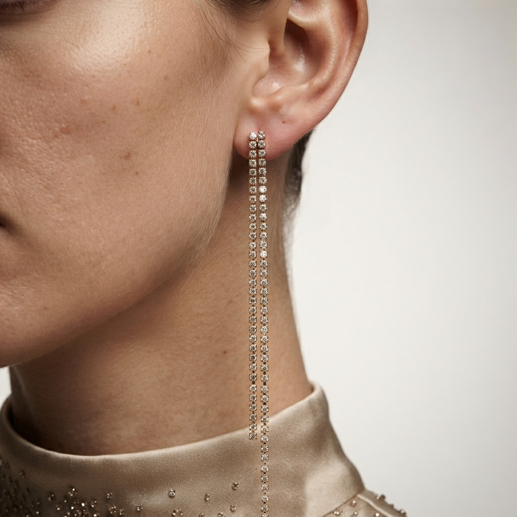 A close-up of a model's ear wearing a long, dangling rhinestone earring with multiple vertical strands of clear stones in a silver-toned setting, paired with a beige satin high-neck top by Civaro