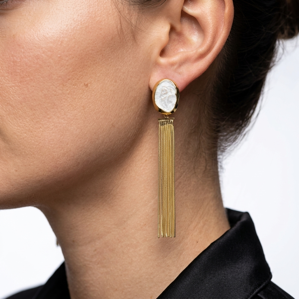 A close-up of a model's ear wearing a gold dangle earring with an oval white cameo-style stone and hanging gold fringe by civaro