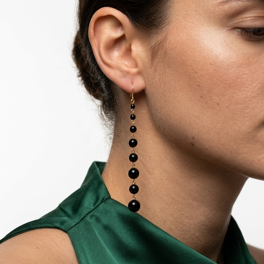 A close-up of a model's ear and neck wearing a long drop earring made of black beads separated by small gold spacers, paired with a dark green satin top against a plain white background, Women's long black pearl earrings with gold tone stainless steel fish hook ear wires designed for weddings, parties and everyday wear by Civaro
