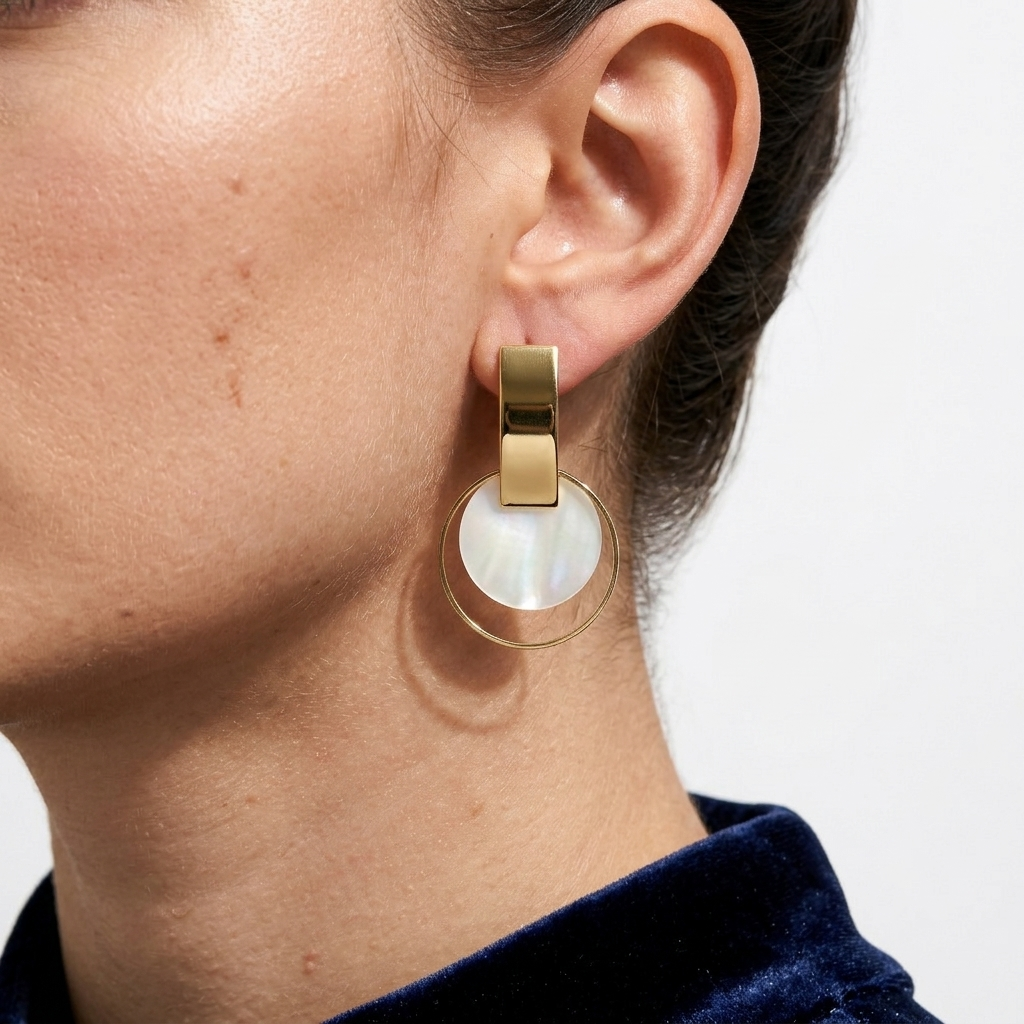 A close-up of a model's ear wearing a gold-tone drop earring with a large circular white shell pendant by Civaro