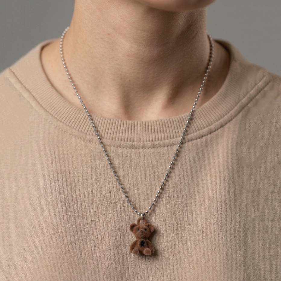 A close-up of a person wearing a silver ball-chain necklace with a small brown teddy bear pendant, paired with a beige crewneck sweatshirt by Civaro